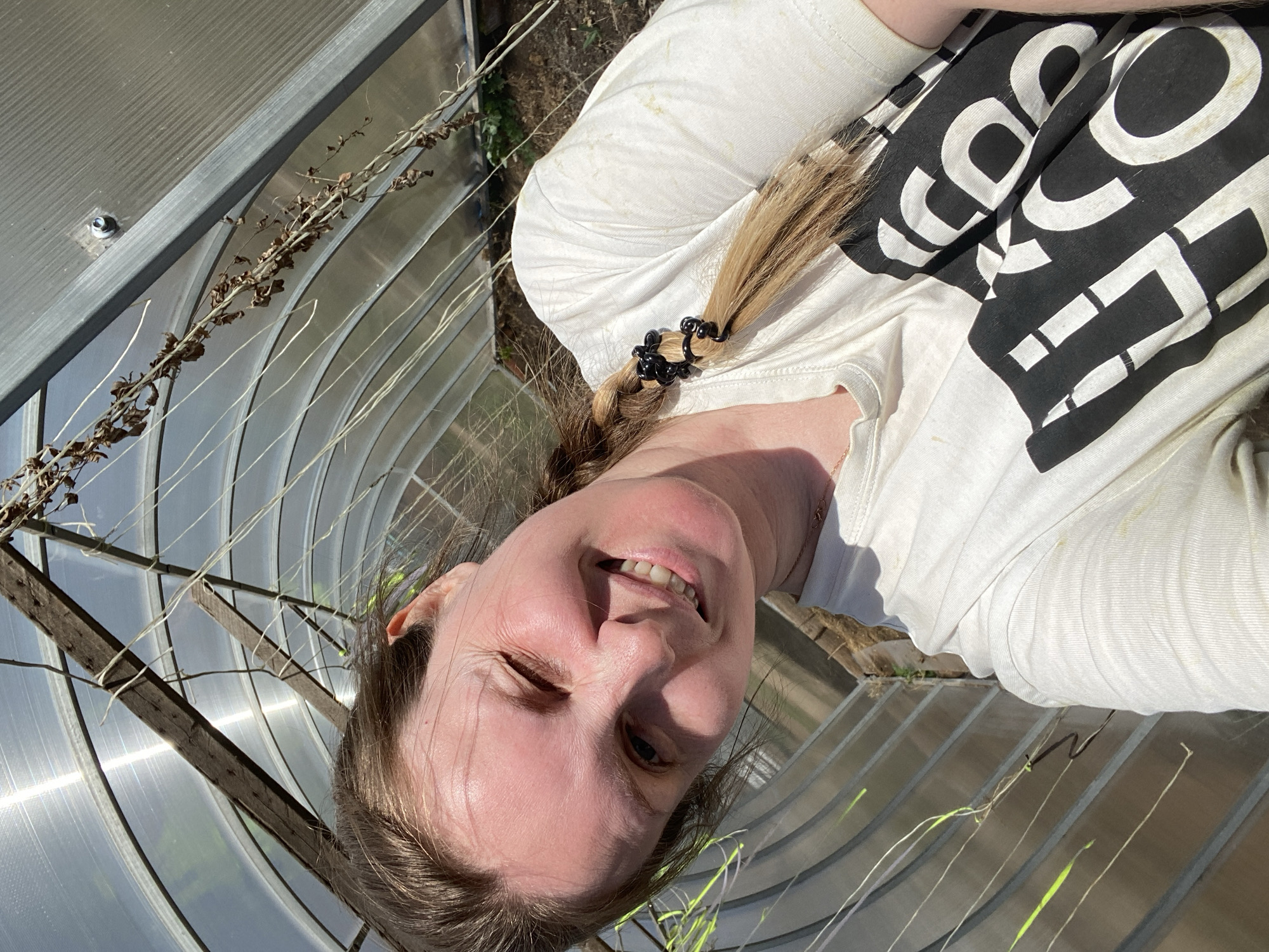 A woman with blonde hair braided and secured with black claw clips smiles at the camera. She is wearing a white t-shirt with black graphic letters. The location appears to be inside a greenhouse with a corrugated plastic roof and wooden supports. Some dried vines and live green plants are visible within the structure. The image lacks specific details that would confirm the location as Chaikovsky, Russia.