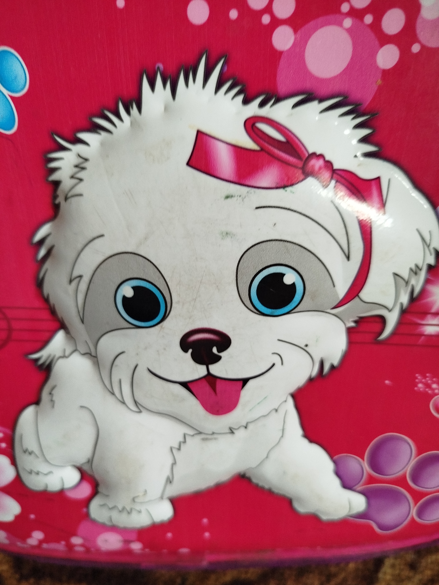 The image displays a close-up, slightly blurred depiction of a cartoon white dog with large blue eyes and a pink bow on its head. The dog is depicted with its mouth open, a pink tongue visible. It has a fluffy white coat and is presented against a bright red background adorned with pink and white circular patterns and some blue petal-like shapes on the left side. There are also purple paw print-like shapes on the right side of the image. The overall impression is of a decorative item, possibly a sticker or part of a child's toy or accessory. The scene is not identifiable as being in Крывасельскі сельскі Савет, Belarus, or any specific urban location.