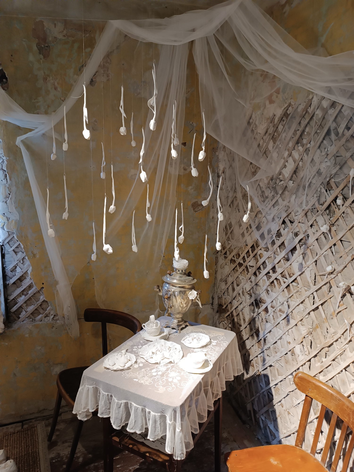 The image shows an interior scene within the city of Yelets, Russia, likely a cafe or art installation. In the center, a small table is draped with a white lace tablecloth adorned with a ruffled edge. On the table, a silver samovar, a teacup, a saucer, and several decorative white plates are arranged. Two wooden chairs are positioned at the table: a darker wooden chair to the left and a lighter wooden chair to the right.
Above the table, white, tulip-shaped objects are suspended from thin wires, creating a decorative canopy. These objects appear to be made of a delicate material and hang at varying lengths, some casting shadows on the wall behind them. The walls of the room are textured and weathered, with patches of peeling yellow paint and exposed plaster and lath. A sheer white fabric is draped across the ceiling and walls, adding to the ethereal and somewhat dilapidated aesthetic of the space. The lighting suggests an indoor setting with natural light filtering in from an unseen source, creating highlights and shadows that emphasize the textures of the environment.