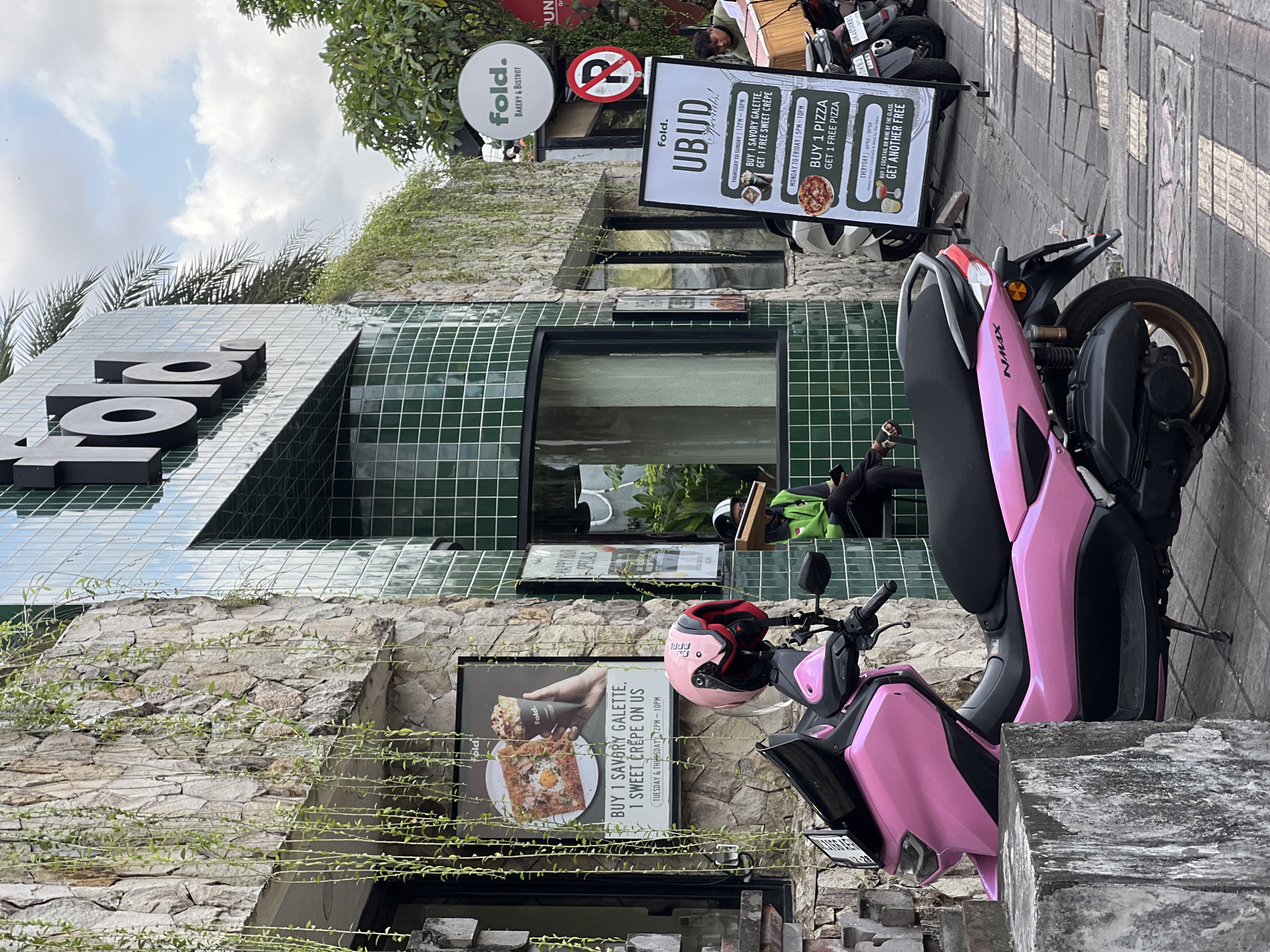 The image shows the exterior of "fold. Bakery & Bistrot" in Ubud District, Indonesia. The building features a stone facade partially covered by climbing plants, a green tiled upper section, and black window frames. Signs advertising the bakery and special offers are displayed, including a "UBUD Specials" sign, and a sign promoting a "Buy 1 Savory Galette, 1 Sweet Crepe on Us" deal. Two parked scooters are visible, one pink and one black. A helmet sits on the pink scooter. Additional signs indicating no parking and the restaurant's name and type are present. A partial view of a person sitting in the background is captured near the black scooter.