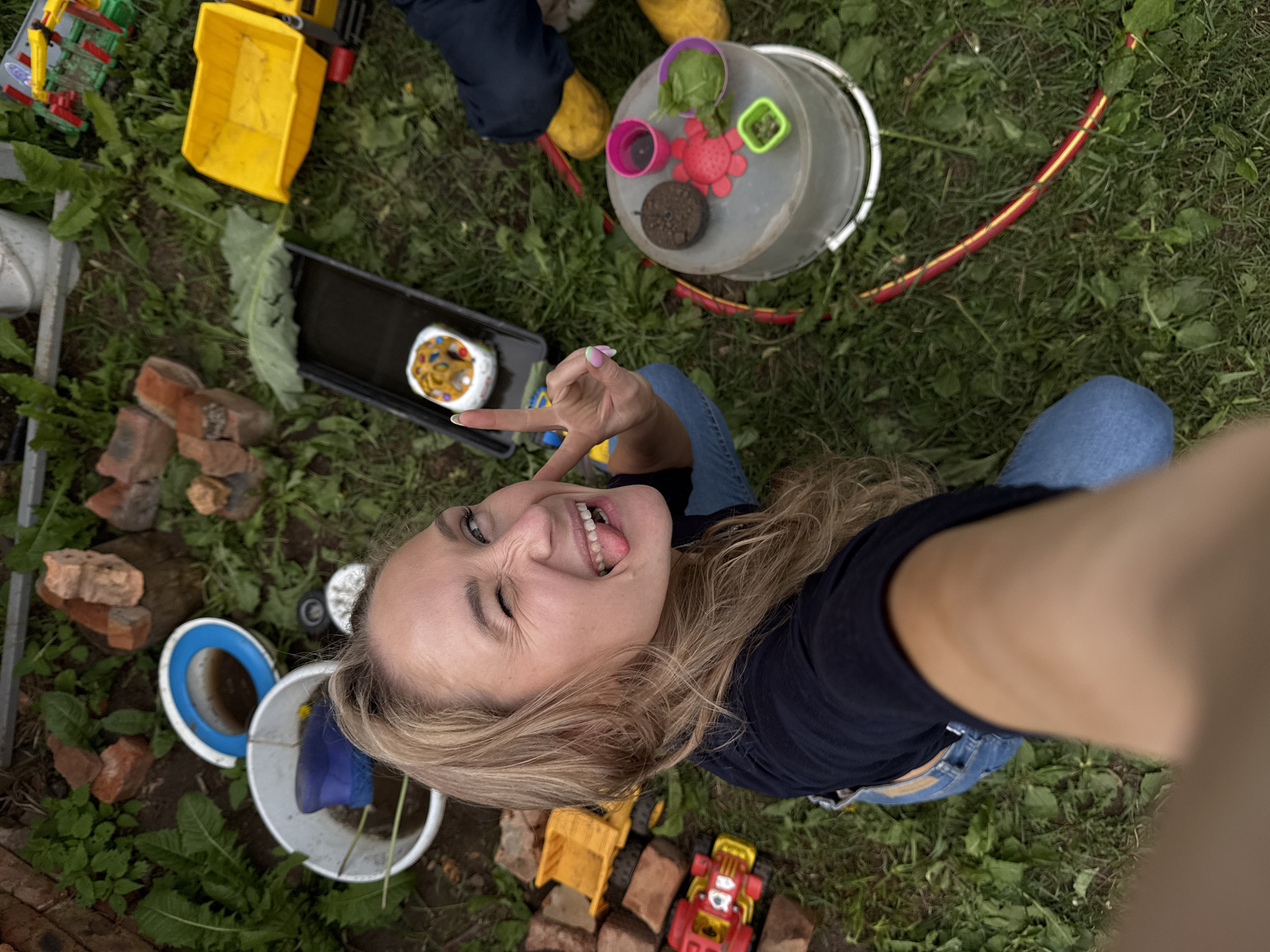 A blonde woman is crouched on the grass in what appears to be a backyard or outdoor play area, possibly in Rosstani, Russia. She is taking a selfie, winking and sticking out her tongue. She is wearing a dark t-shirt and blue jeans. Scattered around her are various children's toys, including a yellow dump truck, a red toy car, a blue bucket with a blue rubber boot inside, and a clear plastic bucket containing small colorful toys and greenery. There are also several bricks and some sort of black tray with a small, decorated object inside. The ground is covered in grass and scattered leaves.