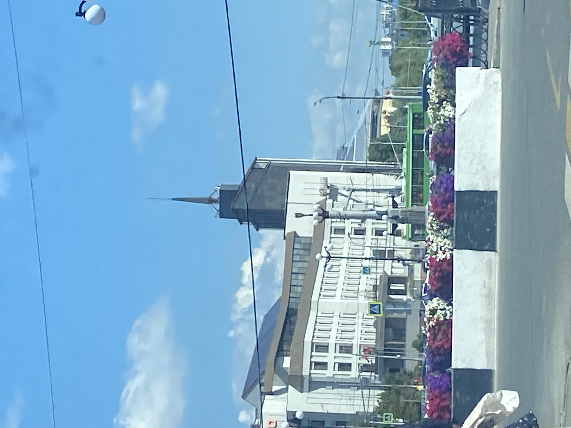 The image shows a street scene in Kazan, Russia. In the background, a modern building with a pointed roof dominates the skyline. In the foreground, there is a row of colorful flowers in planters, and a green bus is visible on the street. The sky is blue with some clouds.