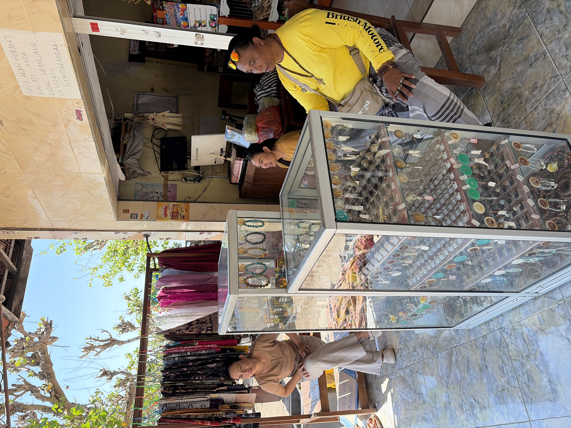The image captures an outdoor market stall in Nusa Penida, Indonesia. In the foreground, two glass display cases filled with various jewelry items are prominent. The case on the right is filled with rings, pendants, and beaded necklaces, while the case on the left displays more necklaces and some bottled items. To the left of the display cases, a woman in a light brown top and beige pants is seated on a wooden bench, looking towards the left. To the right of the display cases, a man wearing a bright yellow long-sleeved shirt, a crossbody bag, and sunglasses is seated on a wooden chair, looking to his left. Behind them, a rack of colorful fabrics hangs, suggesting a clothing section of the market. The background shows a building with a tiled wall and a handwritten sign in Indonesian that reads "SAYA AKAN HORMAT KALAU ANDA TIDAK MEMBUANG SAMPAH SEMBARANGAN" (I will respect you if you do not litter). The overall scene depicts a typical market environment on a sunny day.