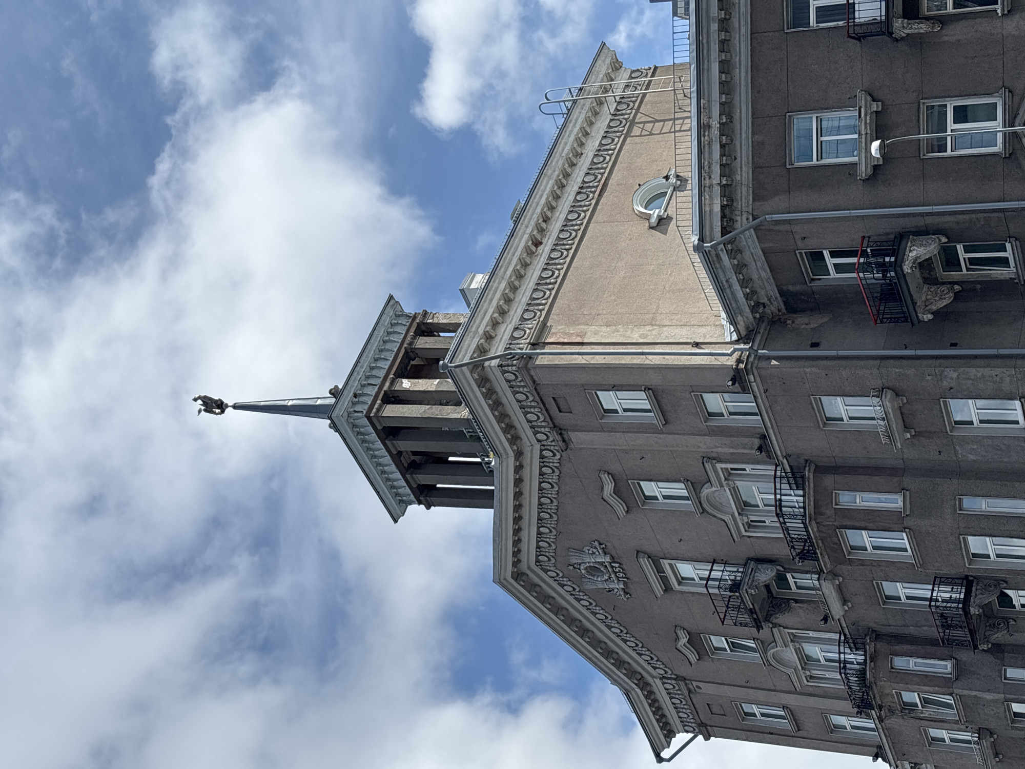 The image shows the corner of a historical building in Tallinn, Estonia, under a partly cloudy sky. The building is a multi-story structure with ornate architectural details, including decorative molding, balconies with intricate metalwork, and a prominent tower topped with a weather vane shaped like a rooster. The facade is a muted brown or grey color. There is a date "1954" visible on a decorative element on the upper part of the building. A fire escape ladder is visible on the side of the building. The image is captured from a low angle, emphasizing the height of the structure against the sky. No people are visible in the image.