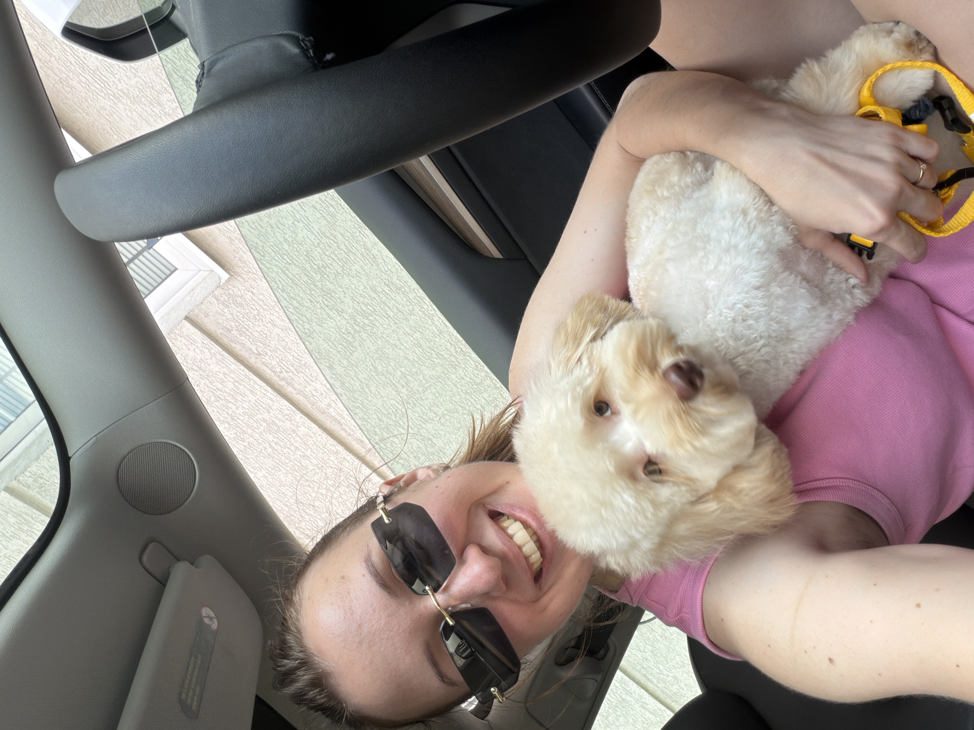 A woman, holding a small, fluffy, light-colored dog, takes a selfie inside a car. The woman is wearing sunglasses and a pink shirt, and she is smiling broadly at the camera. The dog is peeking over her shoulder, also looking towards the camera. The scene appears to be daytime, with natural light illuminating the interior of the car. The background outside the car shows a building with textured siding and windows, suggesting an urban or suburban environment. The interior of the car includes parts of the steering wheel and dashboard, along with a sun visor. The overall mood is happy and relaxed, capturing a moment between a woman and her pet.
