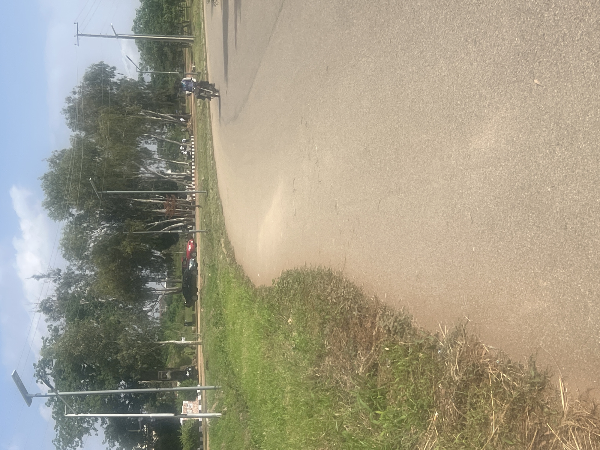This media file captures a scene in Rafin Par, Nigeria, likely during the daytime under a clear, sunny sky, as indicated by the bright lighting and the shadows. The image is oriented vertically, with the asphalt road dominating the right side of the frame. On the left side of the road, a strip of green grass slopes gently upwards, bordered by a line of trees.
In the distance, along the left side of the road, a motorcycle is visible with a rider on it, suggesting movement. Further down the road, a dark-colored car and a red car are parked. Several utility poles with wires are interspersed among the trees, and two solar-powered streetlights are prominent in the foreground, one on the far left and another further back. The environment appears to be rural or semi-rural, characterized by the natural vegetation and the open road. The overall impression is one of a typical day in a Nigerian locale, with everyday activities like road travel and the presence of infrastructure. There is no visible text in the image.