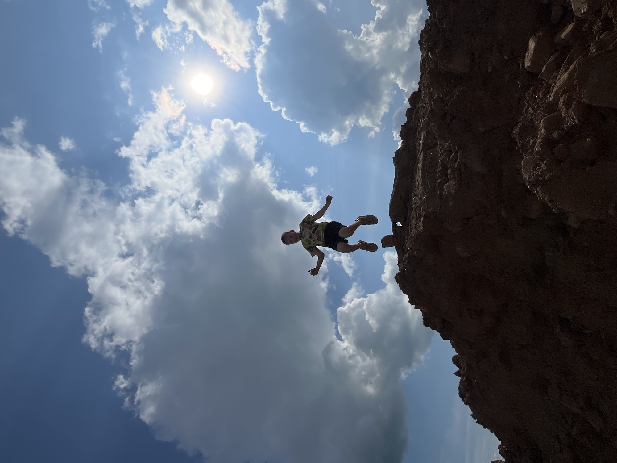A young boy is captured mid-air, jumping over a pile of dark brown dirt. He is wearing a camouflage t-shirt and dark shorts, with his arms raised and legs bent. The sun is bright in the sky, peeking through large, white cumulus clouds. The dirt pile appears to be a large mound of excavated earth, possibly from construction or landscaping. The background is a clear blue sky with the sun. The image is taken from a low angle, emphasizing the boy's jump against the vast sky. The location in the city of Фоки, Russia is not specified within the image itself.