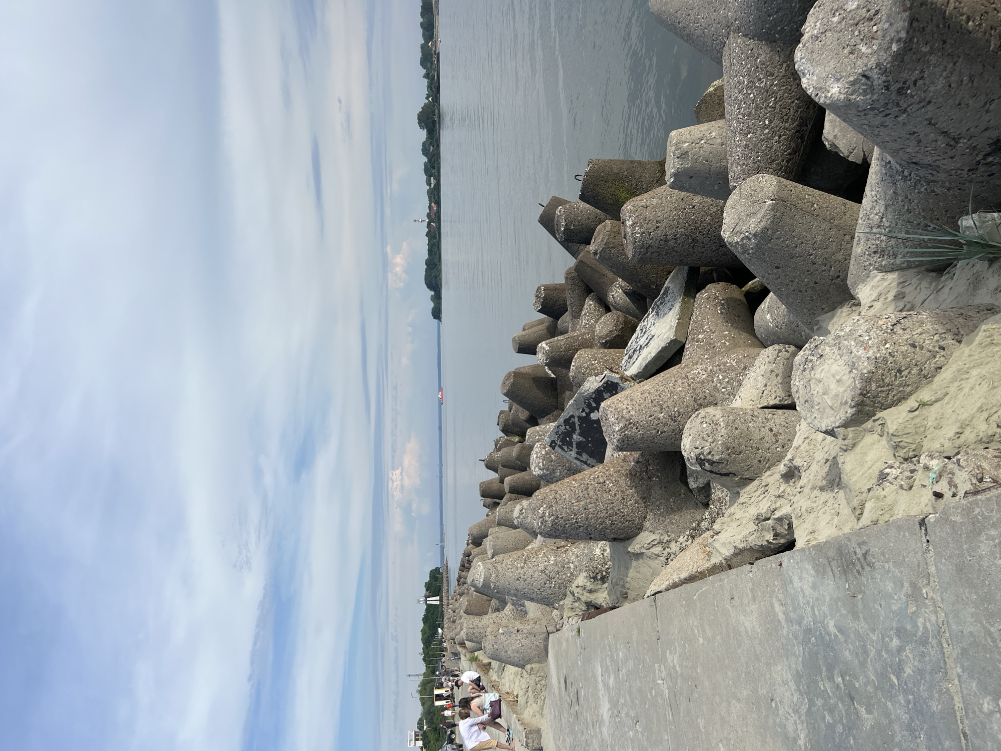 This image depicts a scene in Baltiysk, Russia, featuring a concrete breakwater composed of numerous tetrapods. The breakwater lines the edge of a body of calm water, likely the Baltic Sea. On the left side of the image, a concrete walkway runs parallel to the breakwater, with several people visible walking and sitting on it. Beyond the walkway and breakwater, a coastline with trees and buildings can be seen in the distance, including a prominent lighthouse. The sky is partly cloudy, with a mix of blue sky and white clouds. The overall impression is of a coastal recreational area, possibly a promenade or pier, designed to protect the shore from waves.