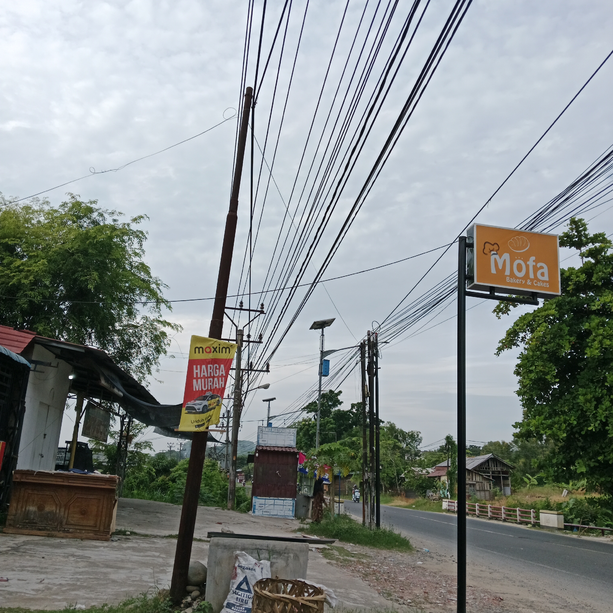 The image depicts a street scene in a rural or suburban area under an overcast sky. In the foreground, a concrete sidewalk runs alongside a paved road. To the left, a wooden counter or display case is partially visible. A large, yellow banner with red text reading "HARGA MURAH" (meaning "cheap price" in Indonesian) and featuring a car graphic is attached to a pole. Above this banner, another smaller sign is visible.
Further down the street, a rectangular orange sign with a chef's hat icon and the text "Mofa Bakery & Cakes" is mounted on a pole. A dense canopy of green trees lines the right side of the road, obscuring some of the background. In the distance, several houses and buildings can be seen, some appearing older or in disrepair. A few utility poles with a complex network of overhead wires are prominent throughout the scene. A solar panel is visible on top of one of the utility poles. A motorcycle is seen in the middle distance traveling on the road. The overall impression is one of a developing area with commercial activity and residential housing coexisting with abundant vegetation.