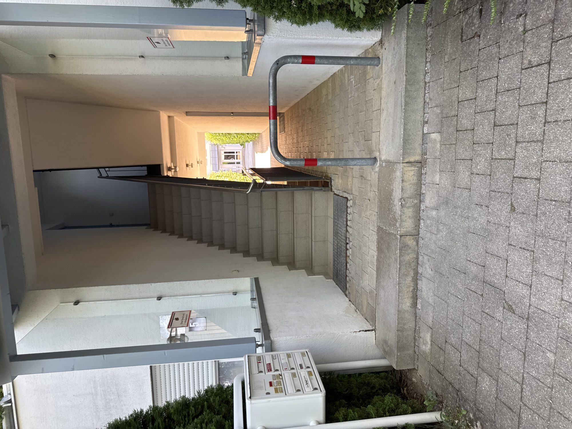 The image shows an exterior view of an apartment building entranceway in Höhenkirchen-Siegertsbrunn, Germany. In the foreground, a gray paved walkway leads up to a concrete step. To the left of the walkway is a white mailbox system with multiple compartments, mounted on white posts, and flanked by dense green coniferous shrubs. A gray metal railing with red reflective markings is positioned across the paved area, just before the stairs.
The central feature is a set of concrete stairs leading upwards into the building. The stairs are covered by a white awning and are bordered by a white wall on the right and a glass partition on the left, which separates the entrance from an exterior area. Beyond the stairs, the hallway is visible, illuminated by overhead lights, and leads to a doorway. The walls of the building are white. On the wall to the right of the stairs, a sign with red lettering and a red border is affixed. Greenery and trees are visible on the left side of the image, and more on the right side further down the hallway. The overall scene appears to be a well-maintained residential building entrance.