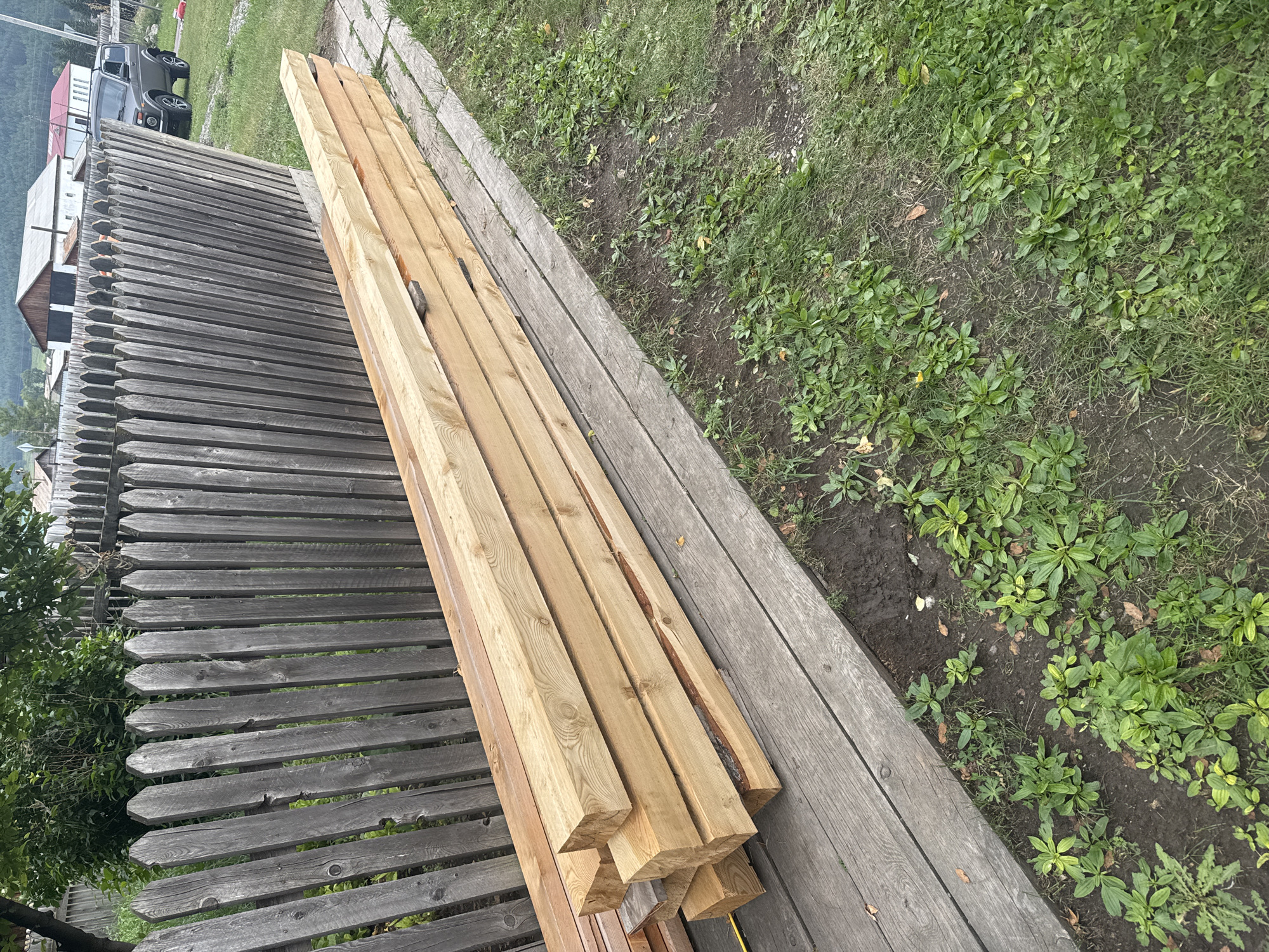 A stack of lumber lies on a wooden walkway next to a weathered wooden fence in Yabogan, Russia. The lumber, comprised of at least nine rectangular beams, is light brown and appears to be new. Behind the fence, trees and buildings can be seen. To the right of the walkway, grass and dirt are visible. A grey SUV is parked on a gravel area to the right of the fence, with buildings in the background.