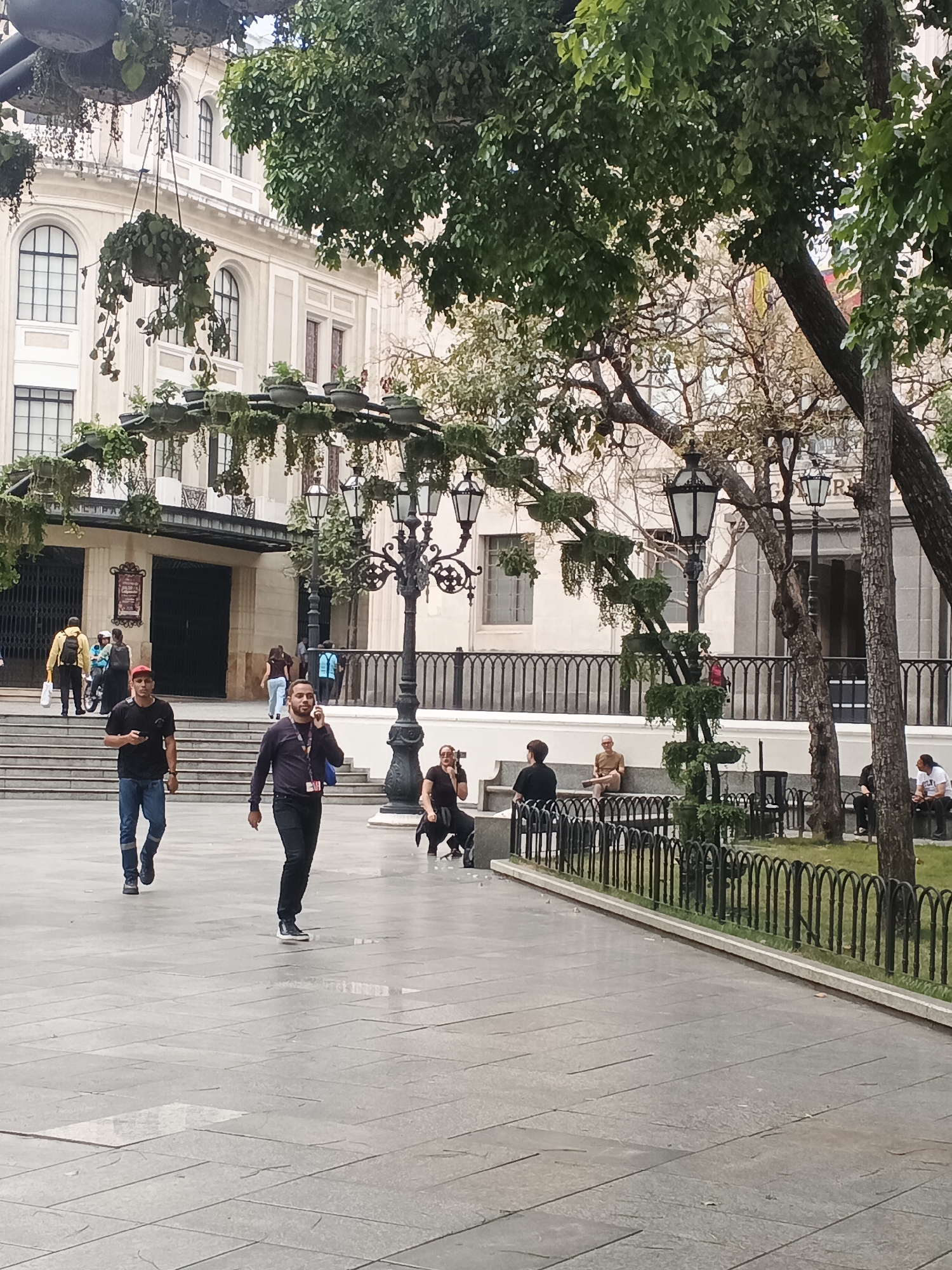 The image captures a bustling public square or park in Caracas, Venezuela, featuring people, lush greenery, and architectural elements under what appears to be an overcast or slightly cloudy day.
In the foreground, two men walk across a light-colored paved area. The man on the left wears a black t-shirt, red cap, and jeans, holding a phone. The man on the right, speaking on a phone, wears a dark shirt and jeans. Further back, several individuals are visible: a person in a yellow jacket and others near steps leading to a large building entrance on the left. On the right side of the frame, several people are seated on a low wall or steps bordering a grassy area. One woman sits on the ground adjusting something.
Dominating the mid-ground are ornate black street lamps and a decorative metal structure adorned with numerous hanging planters and potted plants, creating a canopy of greenery. This structure extends from left to right across the image. The ground is paved with rectangular light gray tiles, some appearing slightly damp.
The background features a grand, light-colored building with classical architectural details, including arched windows and columns. Above its main entrance, partially obscured signs are visible. One sign clearly shows "MUSEO" and another includes the word "BOLIVAR," suggesting it is a museum or historical institution. To the far right background, part of another building is visible, with faint text "P R" on its facade. Large trees with green foliage frame the scene, their branches extending into the upper portions of the image. A low black metal fence separates the paved area from a grassy section of the park on the right.