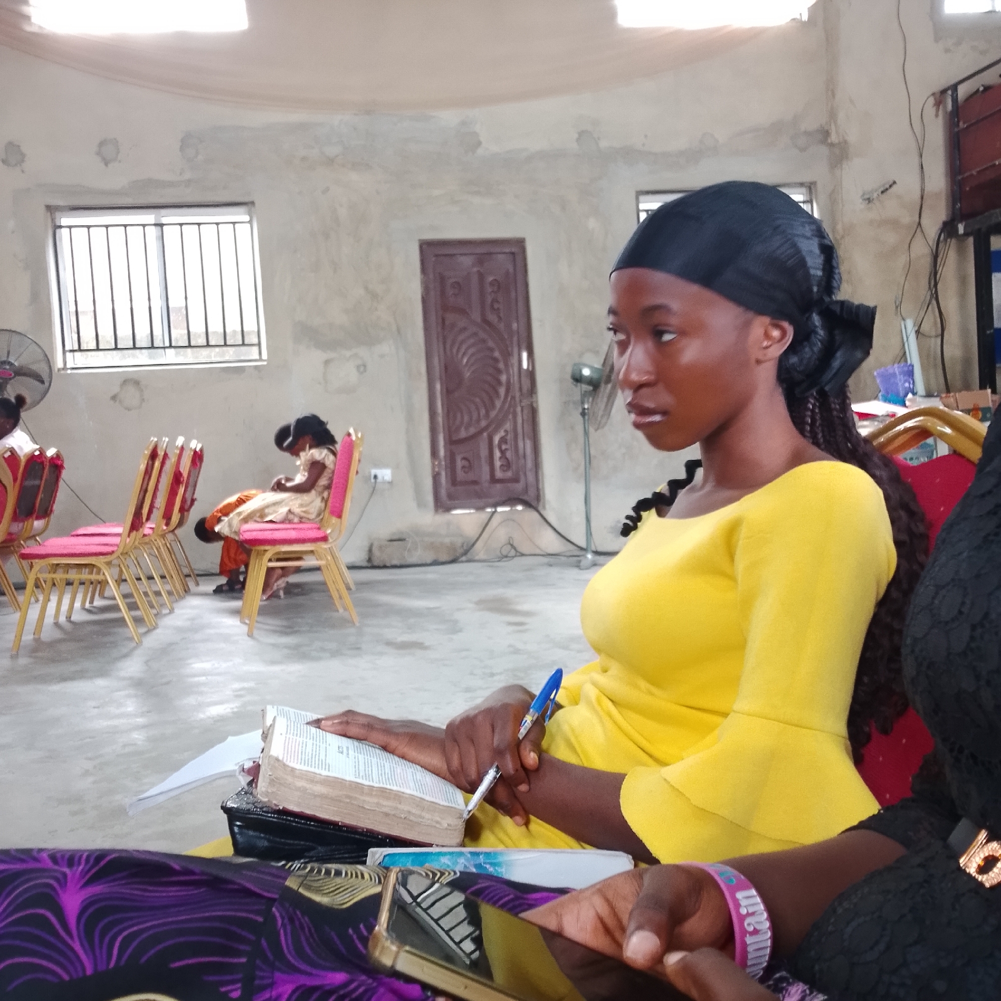 In Ado, Nigeria, a young woman with dark skin and braided hair, wearing a black headscarf and a bright yellow dress, is seated. She is focused on a bible that lies open on her lap, a pen in her hand, suggesting she is reading or studying. The setting appears to be a room with a concrete floor, light-colored walls, and a high ceiling with a visible cloth covering. There are windows with metal bars. Other people are visible in the background, seated on chairs, and a fan is present. The overall impression is of a communal gathering or service.