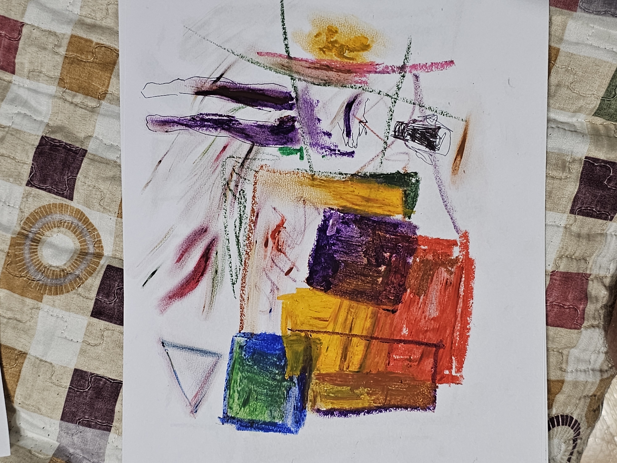 The image depicts an abstract drawing on a white piece of paper. The drawing features a combination of geometric shapes and loose lines created with what appears to be oil pastels or crayons. Colors include blue, green, yellow, orange, red, purple, brown, and pink. The shapes are arranged in a somewhat haphazard manner, with some overlapping and others appearing as isolated elements. The background is blurred, revealing a patterned fabric with squares in various earth tones. The drawing's subject is unknown. There is no indication of location, so I cannot associate it with Plovdiv, Bulgaria.