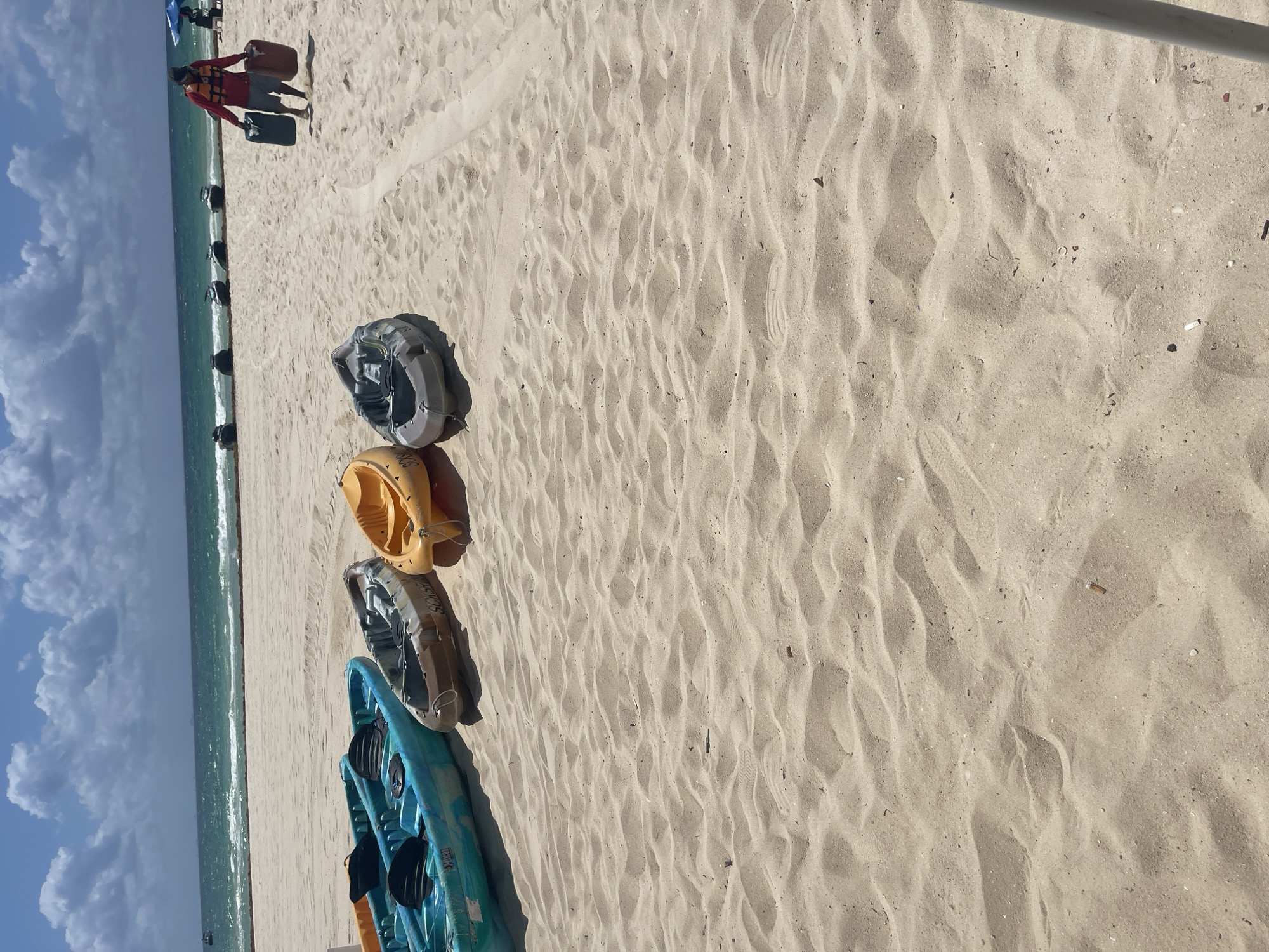 The scene is a beach in Playa del Carmen, Mexico. The sky is partially cloudy with the ocean visible on the left side of the frame. Four kayaks, grey, yellow, beige, and blue are resting on the light colored sand. An individual, in the upper left corner, is carrying two black containers, and is wearing a red shirt and orange life vest.