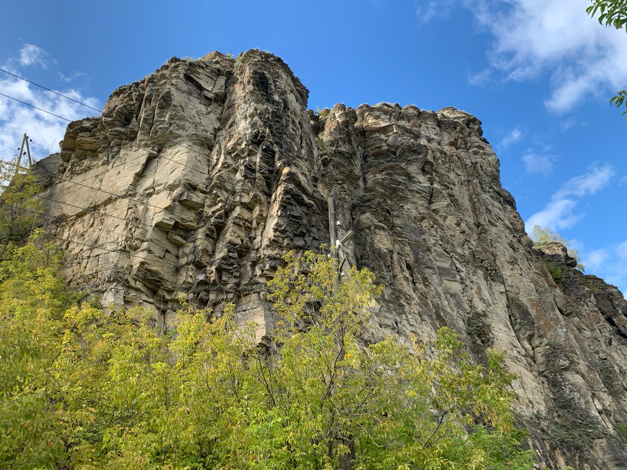 The image shows a large rock cliff face with a blue sky and white clouds above. In the foreground, there is green foliage from trees. Power lines are visible running across the cliff face. A wooden utility pole with some fixtures is also present, attached to the cliff. The rock face is layered and textured, with areas of dark and light rock, and some patches of moss or lichen. The scene appears to be in an outdoor, natural environment, possibly at the edge of a city or town. Based on the provided information, this location is within the city of Minyar, Russia.