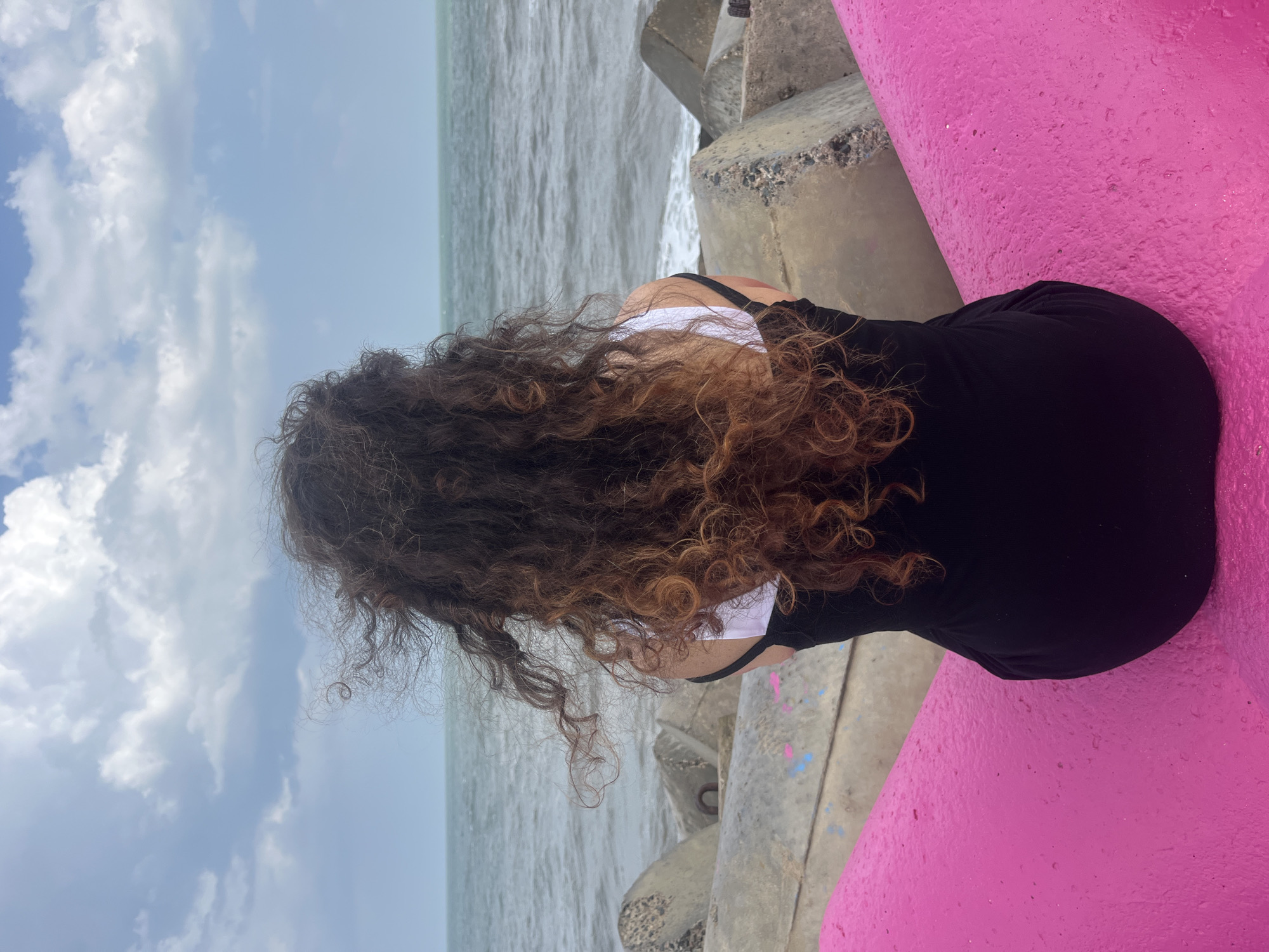 A woman with long, curly brown hair is seated facing away from the camera, looking out at a choppy grey sea under a cloudy sky. She is wearing a black dress with a white strap visible. The woman is positioned on a bright pink, curved structure, which appears to be part of a public seating area or art installation. Behind her, grey concrete tetrapods line the shore, serving as a barrier against the waves. The sea is a deep grey, with white foam crashing against the tetrapods. The sky is filled with dynamic, textured clouds in shades of white and grey, with patches of lighter blue peeking through. The overall impression is one of a person contemplating the vastness of the ocean. This scene is not located in the city of Phú Thủy, Vietnam.