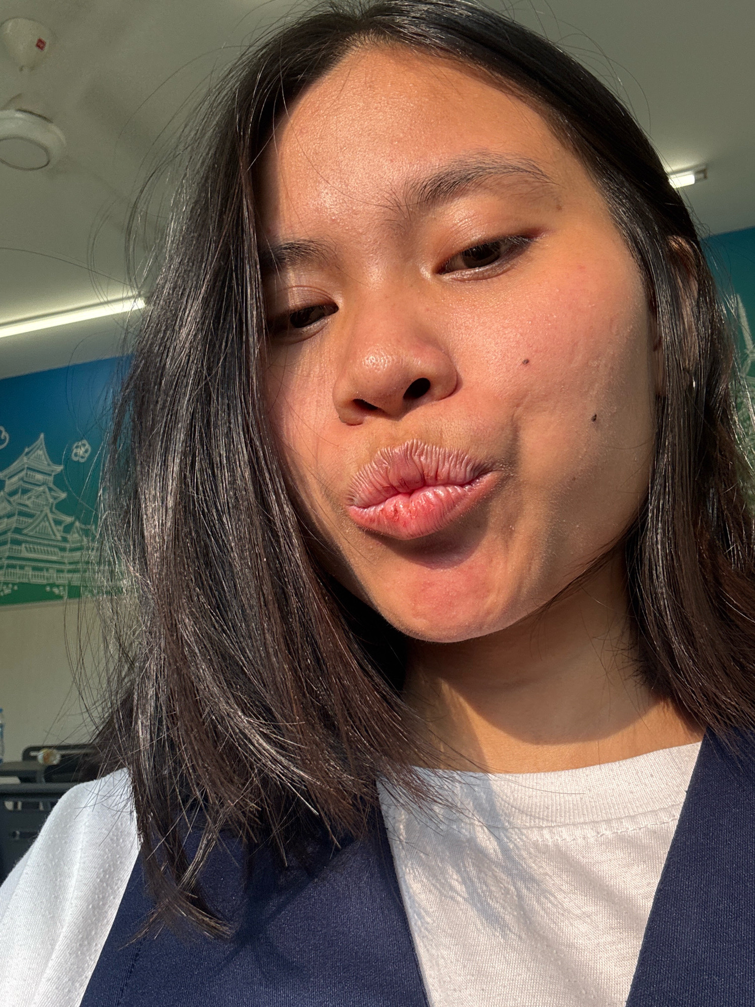 In a close-up, low-angle selfie, a young East Asian woman with dark hair styled down her shoulders pouts her lips. She is wearing a white t-shirt and a dark blue vest. The background shows a mural with a white castle on a blue and green gradient, and a white ceiling with a smoke detector. The lighting suggests it is daytime, with sunlight casting shadows on her face and clothing. The location appears to be indoors, possibly a public space or commercial establishment, given the mural and ceiling fixtures.