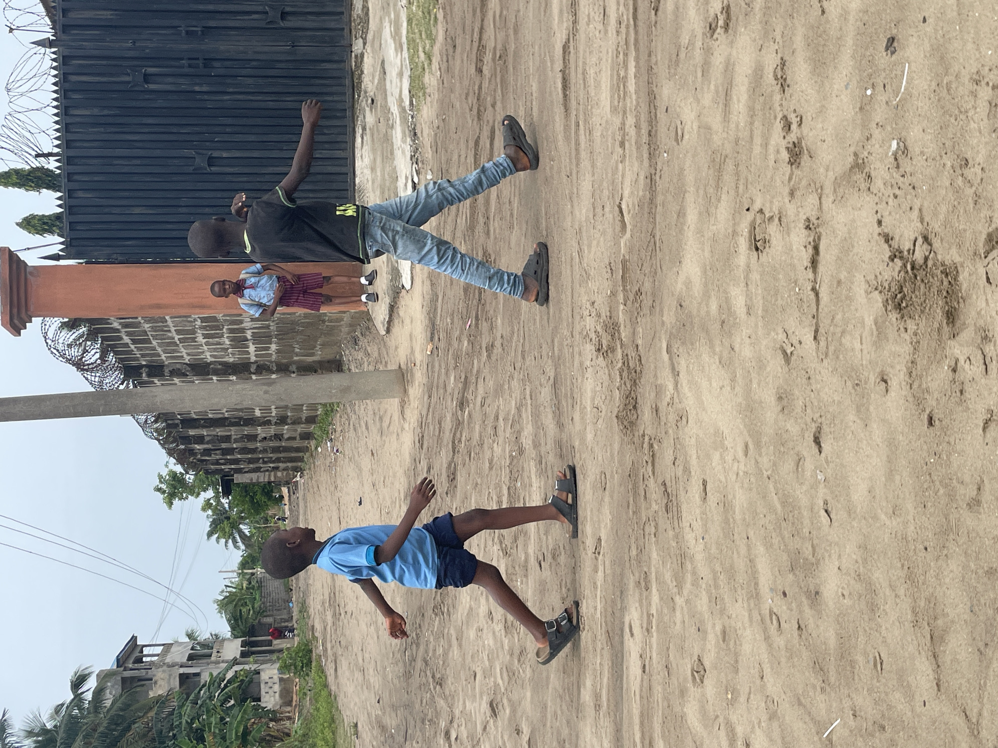 The scene depicts a sandy street in Badagry, Nigeria. Two boys are in motion, one running in a blue shirt and dark shorts, and another walking with arms outstretched wearing jeans and a black T-shirt. A third child in a school uniform stands near a gate. The environment includes concrete buildings, a black metal gate with barbed wire, utility poles, trees, and a sandy ground covering the street.