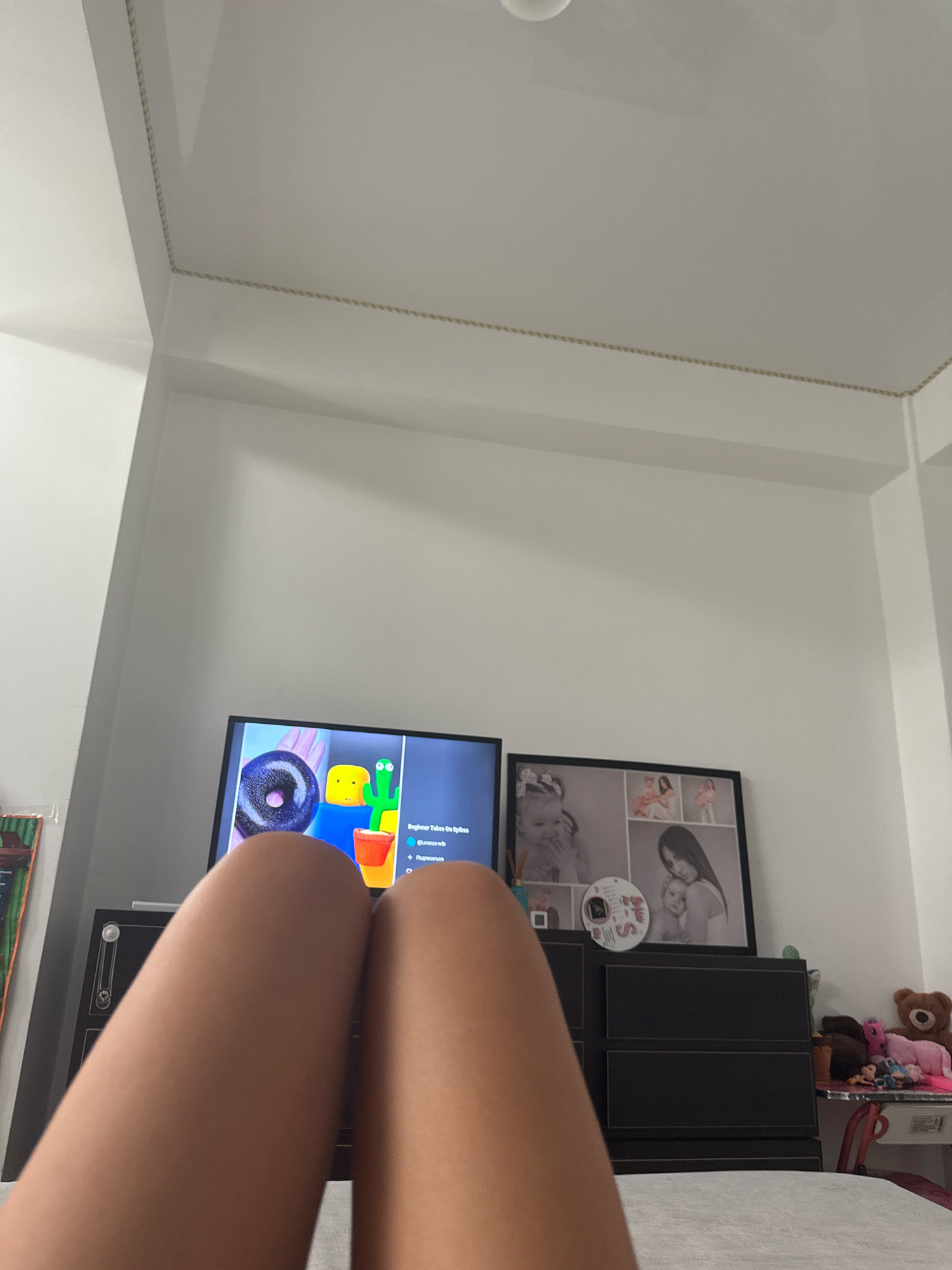 This is a low-angle, indoor, point-of-view shot of a person's legs in the foreground, with a television screen, a framed collage of photos, and a dresser with various objects on and around it in the background. The person is lying down, with their legs extended towards the camera. The television screen displays a video with animated characters and text. The photo collage on the wall contains several pictures of a woman and children. The dresser is dark-colored, and on it are a teddy bear, stuffed animals, and other small items. The overall impression is of a bedroom or living space. The image does not provide any specific geographical information about Chișinău, Moldova.