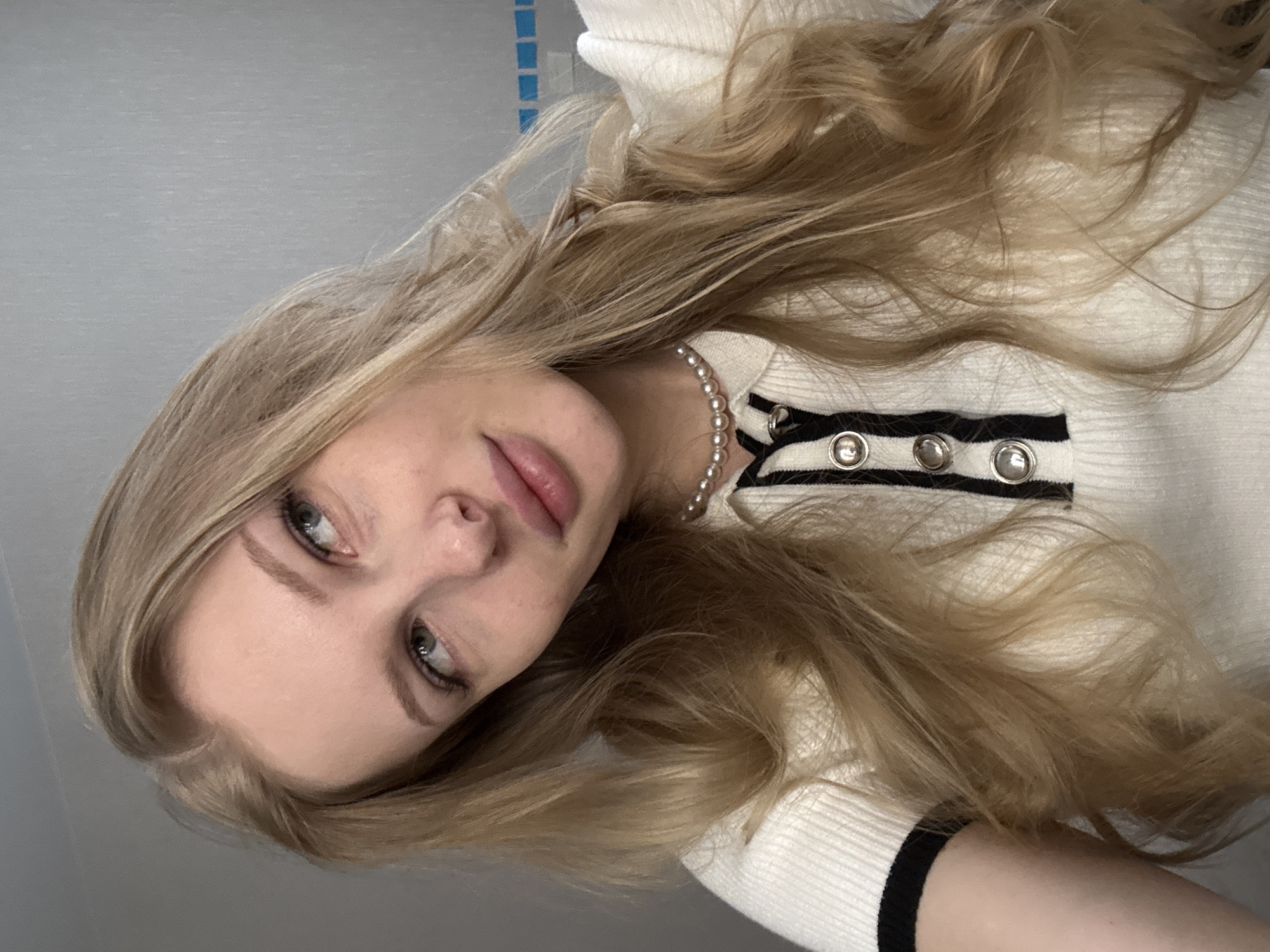 A young adult female with long, wavy, light-blonde hair, parted on the viewer's left, is depicted in a close-up, chest-up portrait. Her head is tilted, and her light-colored eyes are directed upwards and to her right, not meeting the camera. Her lips are naturally full and slightly parted. She wears an off-white, ribbed knit polo-style shirt featuring a black vertical placket with three prominent silver domed buttons and a black trim on the short sleeve visible on her left arm. A single strand of small, round, white pearls is worn as a choker necklace. The background is a plain, light grey wall, with faint horizontal blue lines visible on the far right side, possibly indicative of a bathroom or tiled area. The scene is situated in Perm, Russia.