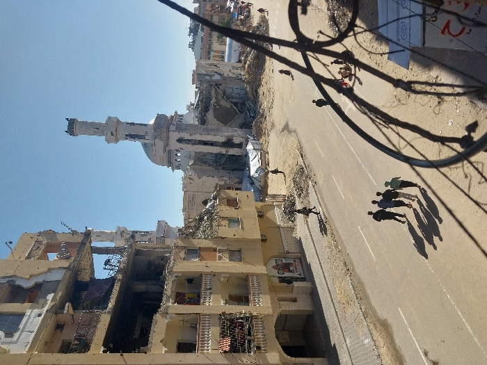 This image depicts a scene of extensive structural damage in the city of Khan Yunis, Palestinian Territory, under a clear blue sky. In the foreground, out-of-focus dark wires or cables partially obscure the lower right portion of the view.
The central feature is a wide asphalt road, showing white double solid and single broken lane markings, with dirt and debris accumulated along its edges. Four males in dark clothing walk in a single file line across the road from left to right, casting long shadows ahead of them. Further down the road, on the right, a small group of approximately 6-8 individuals, including children, are gathered or seated amidst rubble. Several other pedestrians are visible on sidewalks and dirt paths throughout the scene.
To the left of the road stands a multi-story residential or commercial building, exhibiting severe structural damage with large sections of its facade and internal floors collapsed or exposed. Balconies are damaged, and debris is evident around its base. A few individuals walk on the sidewalk directly adjacent to this damaged building.
In the mid-ground and background, the dominant structure is a large mosque with a prominent minaret and dome, both significantly damaged. The minaret displays pockmarks and missing sections, while parts of the dome and surrounding mosque complex are visibly destroyed and reduced to rubble. Beyond the mosque, additional urban buildings are visible. To the right of the road, an open, unpaved area strewn with debris leads to several white tarpaulin shelters or tents in the distance. The scene is illuminated by strong sunlight, creating distinct shadows.