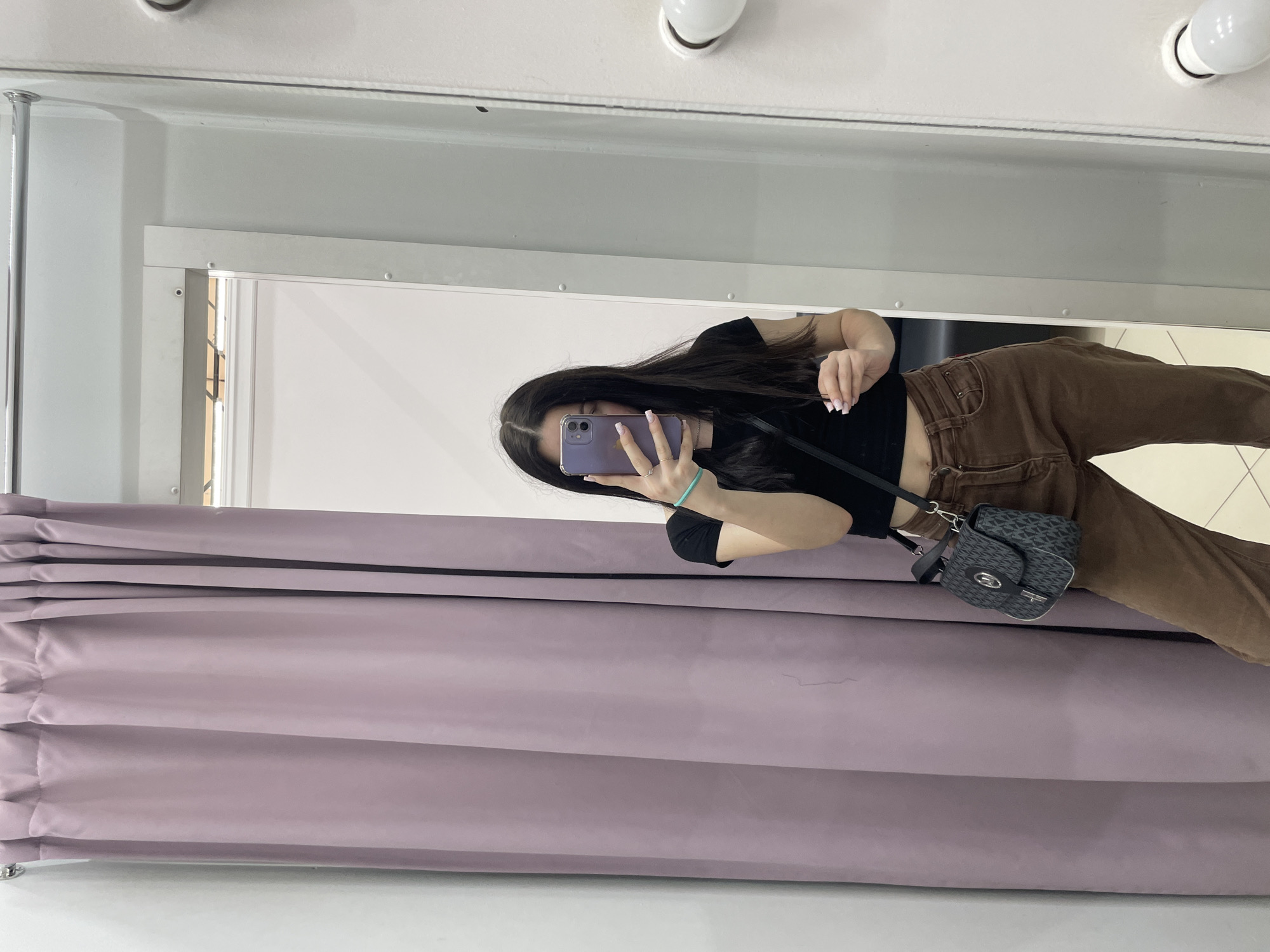 A young woman with long dark hair takes a selfie in a fitting room. She is wearing a black crop top, brown pants, and a small patterned crossbody bag. She has a light blue bracelet on her left wrist and is holding her phone, which has a purple case. The background consists of a pale purple curtain on the left and a mirror on the right, which reflects part of the fitting room and a light fixture. The floor appears to be tiled. The image does not contain any specific indicators to determine its location within Chaikovsky, Russia.