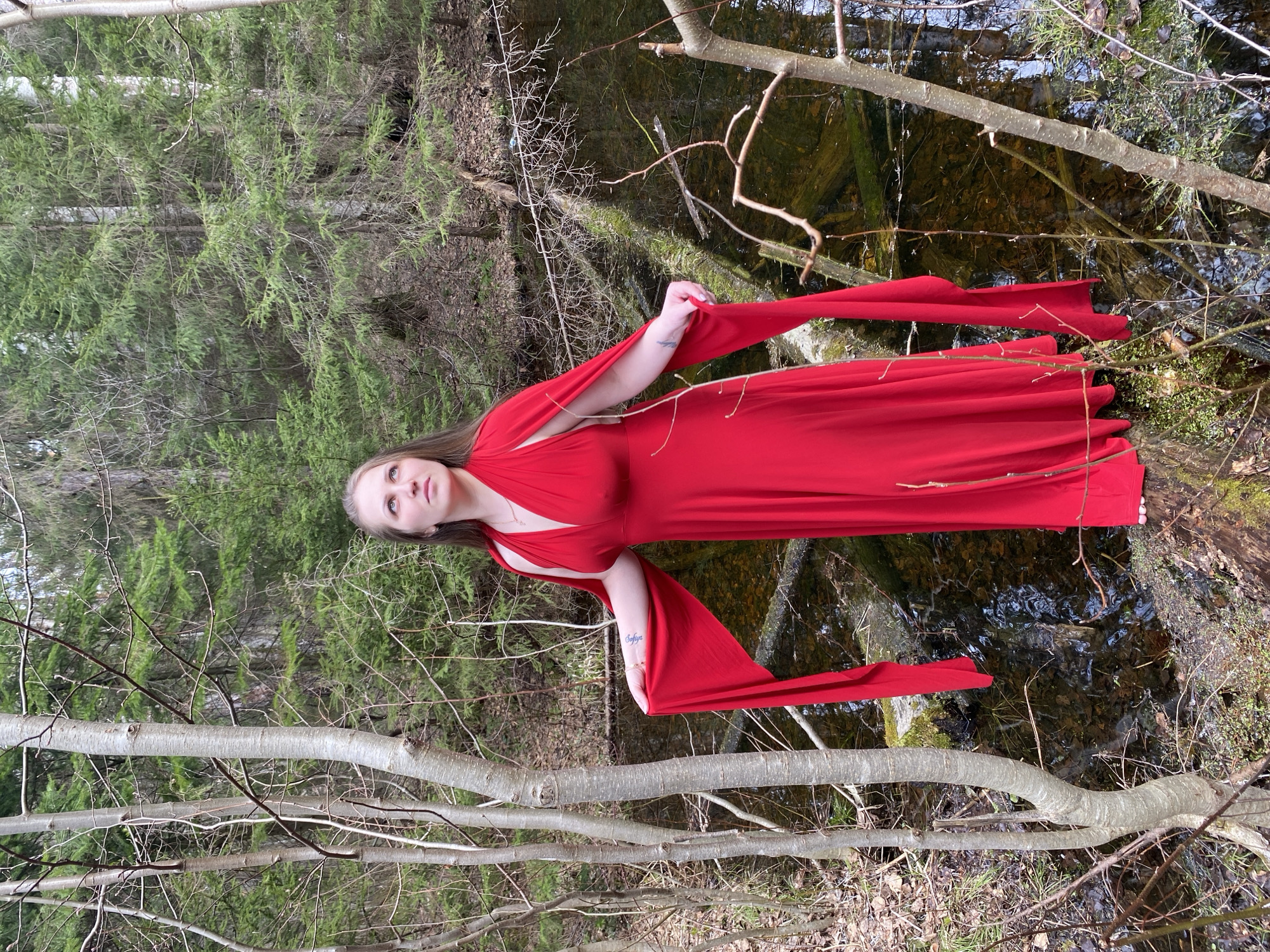 A young woman with long, light brown hair is positioned in a natural, forested setting. She is wearing a flowing, vibrant red dress with long, draping fabric elements, some of which extend into a calm, dark body of water beside her. Her gaze is directed upwards and slightly away from the viewer, conveying a serene or contemplative expression.
The background features a dense growth of dark green evergreen trees. In the foreground and midground, several bare, light-colored tree branches, ranging from thick limbs to thin twigs, crisscross the scene horizontally, partially framing the subject. The body of water, possibly a shallow stream or pond, shows reflections of the environment and reveals submerged debris like branches, leaves, and moss. Patches of dry leaves and sparse ground cover are visible on the banks.
The lighting is soft and diffused, indicating daytime conditions without direct harsh sunlight. On the woman's left forearm, a script tattoo is visible, spelling the name "Sofiya." The striking red dress provides a vivid contrast against the muted greens, browns, and grays of the surrounding forest and water. The overall scene suggests an artistic portrait session within a temperate woodland environment, consistent with the Марковский, Russia location.
