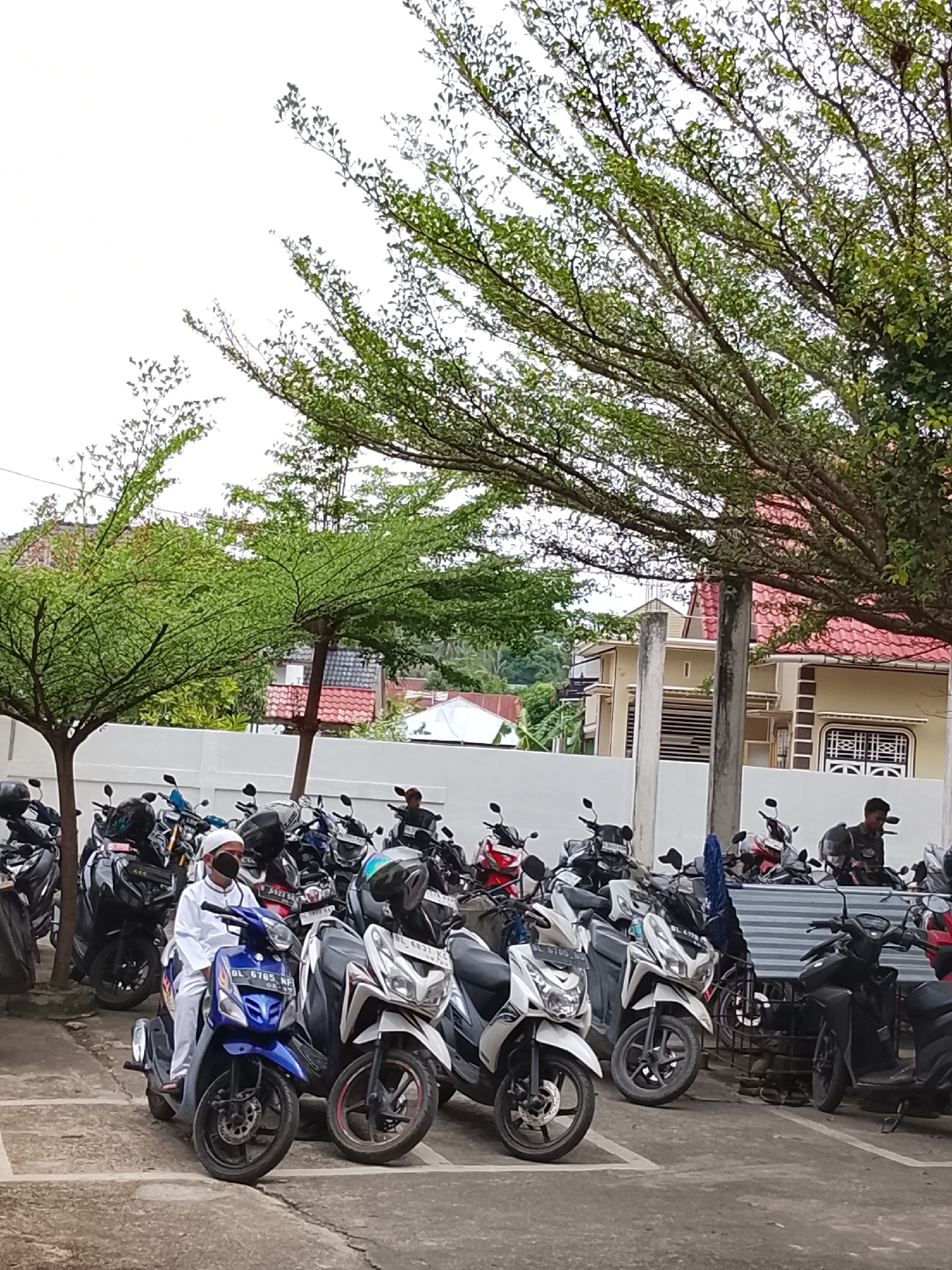 In Lhokseumawe, Indonesia, a man in white Islamic attire and a black mask is seated on a blue scooter. The scooter, bearing the license plate DL 6785 MF, is parked among a row of other scooters. The background features a white wall, trees with sparse green leaves, and buildings with red-tiled roofs. The sky is overcast. The scene appears to be an outdoor parking lot.