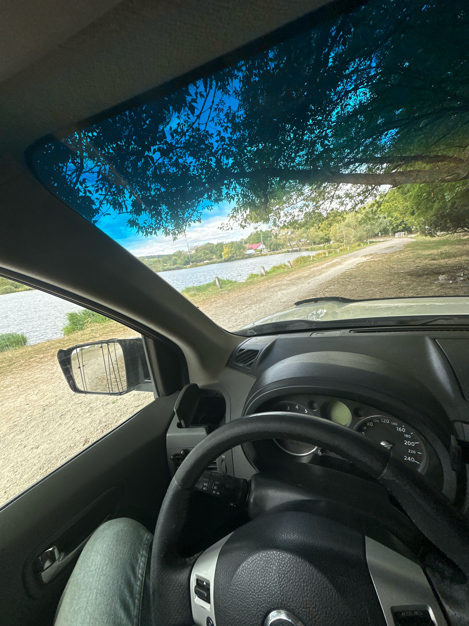 This is a point-of-view shot from inside a car, looking forward and slightly to the left. The car is parked on a dirt road next to a body of water. The windshield is visible, with tree branches and blue sky reflected on it. Through the windshield, a lake or river is visible, with a small building on the far bank and trees lining the horizon. The dashboard and steering wheel of the car are in the foreground, with a leg clad in light-colored jeans visible on the left. The scene appears to be in a rural or semi-rural setting. No specific location in Makiv, Ukraine is identifiable from the image.