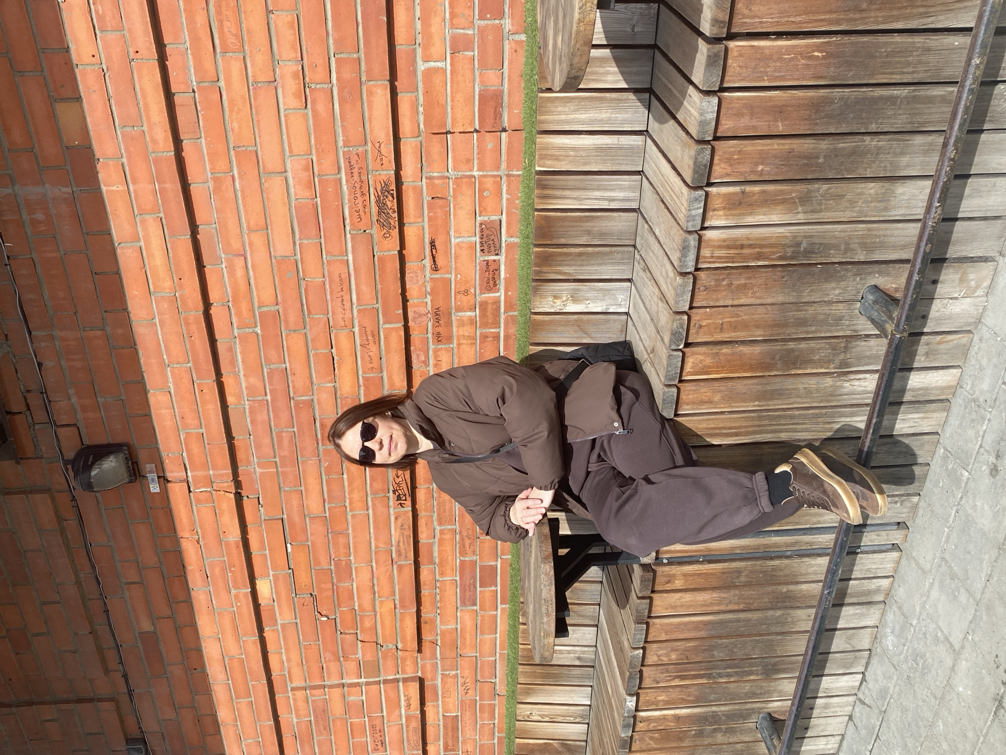 A person, presumably a woman, is sitting on a wooden bench or platform. She is wearing sunglasses, a brown puffer jacket, and matching brown pants and shoes. She has her hands clasped in front of her.
She is positioned against a brick wall that is partially covered in graffiti. The brick wall appears to be weathered and has some electrical fixtures attached. Next to the platform is a patch of green, potentially grass or artificial turf. There is a metal railing next to the seating area. The ground surrounding the area appears to be concrete.
Based on the graffiti and the image's association with Yekaterinburg, Russia, it is likely the image was taken in a public space within the city.