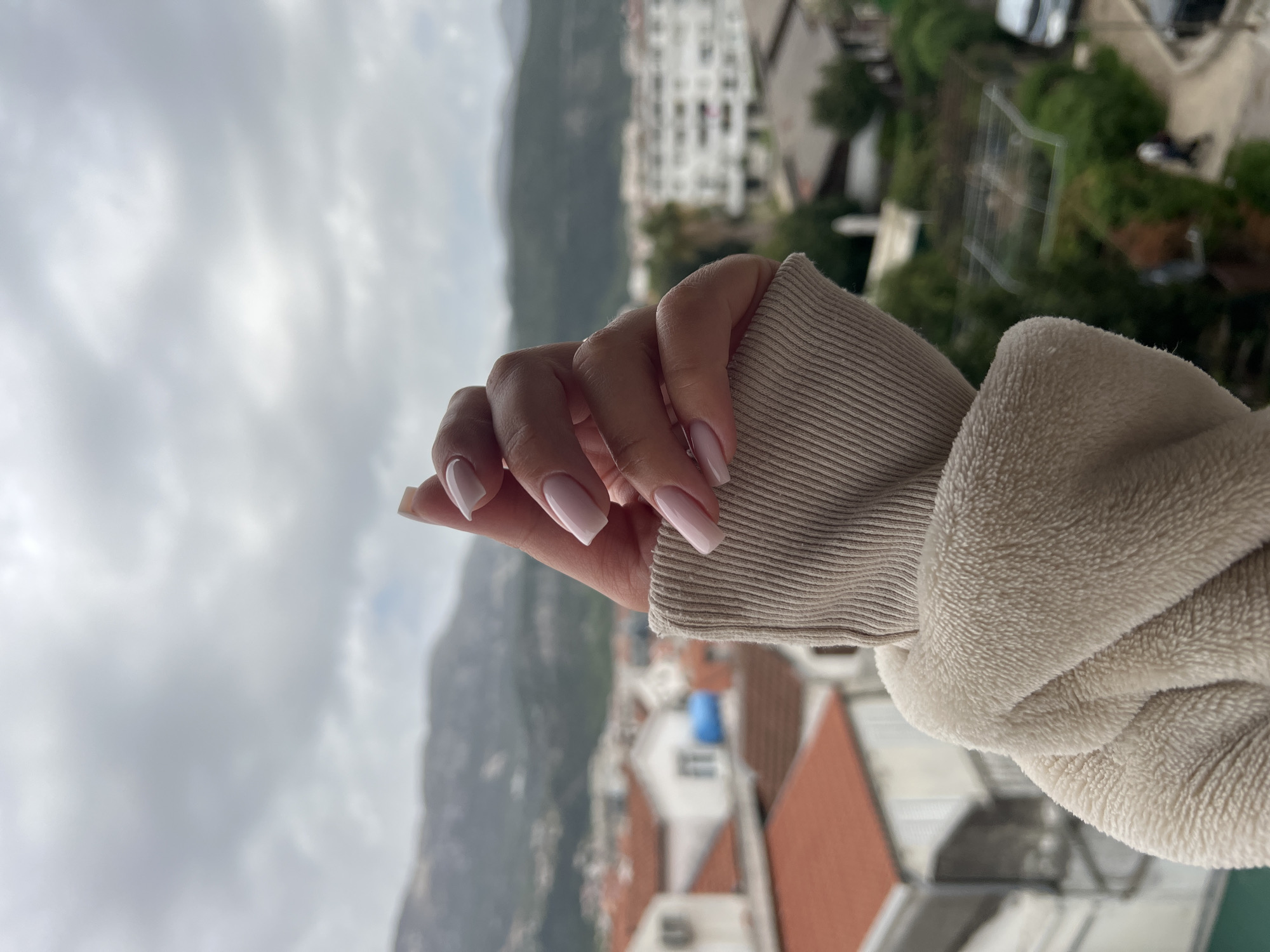 A hand with manicured nails is reaching out in front of a cityscape. The hand is wearing a light beige ribbed sweater sleeve. The fingernails are square-shaped with a light pink base and white tips. The background shows a view of Budva, Montenegro, with mountains, buildings, and a cloudy sky. Red rooftops and green trees are visible among the buildings.