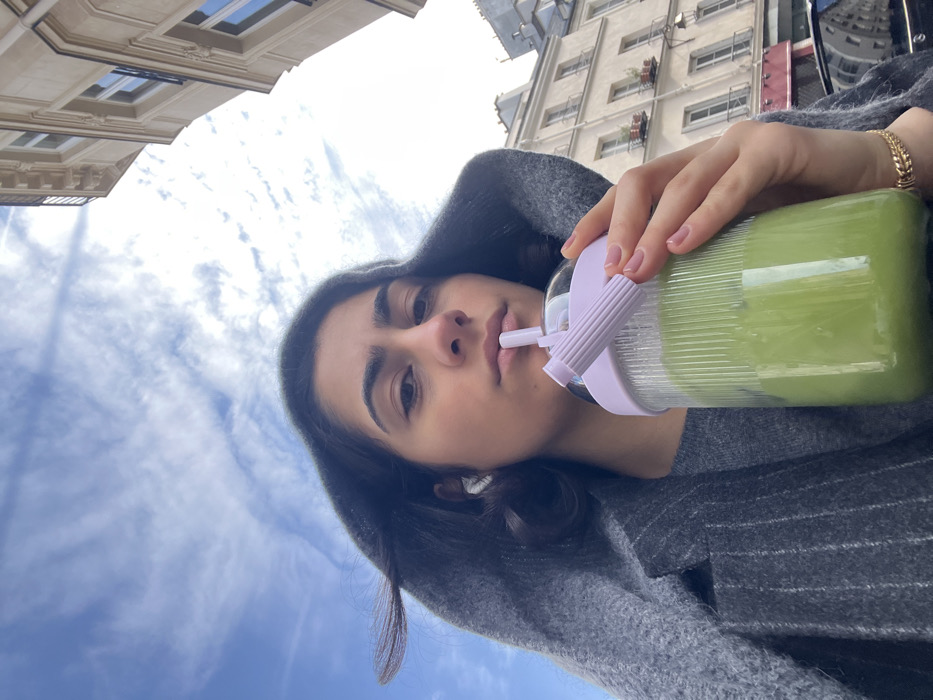 A young woman is captured in a low-angle outdoor portrait, positioned to consume a bright green liquid from a transparent plastic tumbler through a straw. She has dark hair, partially covered by a grey hooded garment that is pulled over her head. She looks directly forward, and white wireless earbuds are present in both ears. Beneath the hooded layer, a grey pinstripe jacket is discernible. A gold-toned chain link bracelet is visible on her left wrist, and her fingernails are painted with a clear or light pink polish. The tumbler features a light purple lid with a fixed straw. The background consists of a blue sky with scattered white clouds. To the right, a multi-story building of classic Haussmannian architecture is present, identifiable by its light-colored stone facade, regular window patterns, and decorative cornices and balconies. The scene takes place on a street within the city of Paris, France.