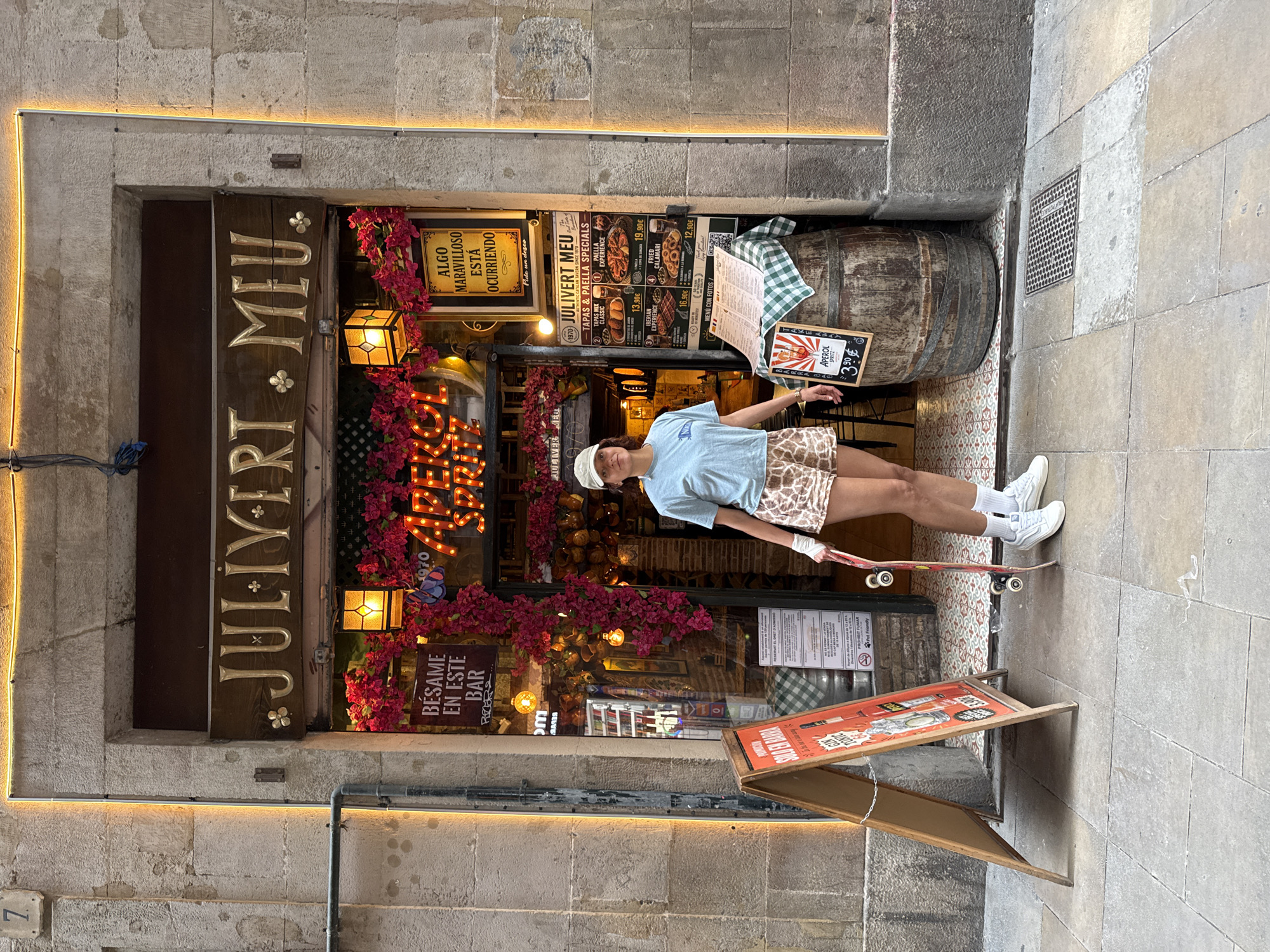 Here is a description of the image:
The photograph captures a scene in front of a restaurant named "Julivert Meu" in Barcelona, Spain. The restaurant's facade is adorned with warm lighting, flowers, and signs in Spanish. There's a wooden barrel situated next to the entrance, and a sandwich board stands on the sidewalk, showcasing the menu. A young woman stands with a skateboard, leaning against the entrance. She is wearing a light blue t-shirt, patterned shorts, white sneakers, and a white baseball cap. The setting is an urban street, with stone walls and pavement visible in the background.