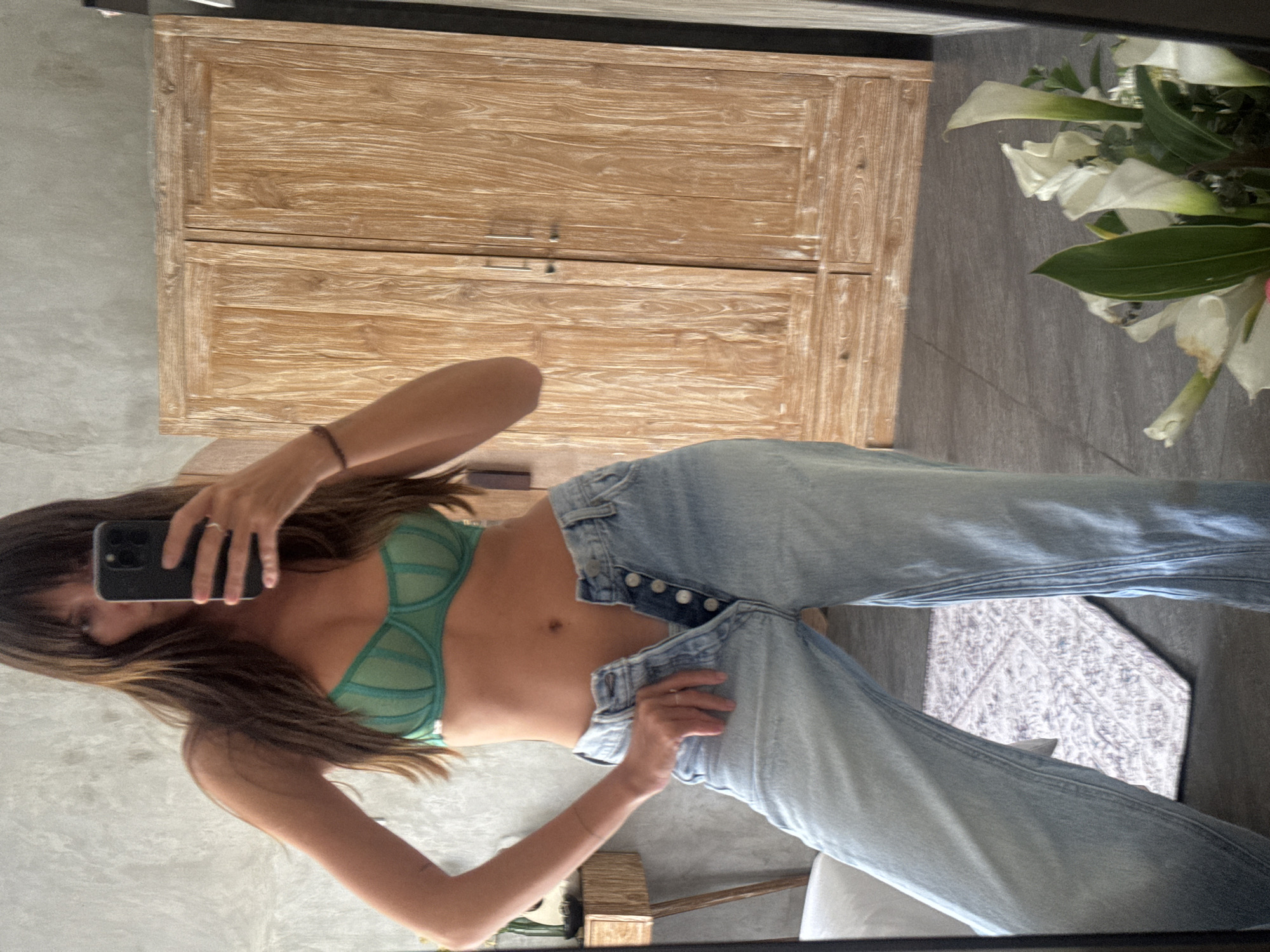 A woman with brown hair is taking a selfie in front of a mirror. She is wearing a green bra and unbuttoned light blue jeans. Behind her is a light-colored wooden cabinet and a vase of white calla lilies. A rug with a geometric pattern is visible on the floor. The location is inferred to be North Kuta, Indonesia, based on the user's prompt.