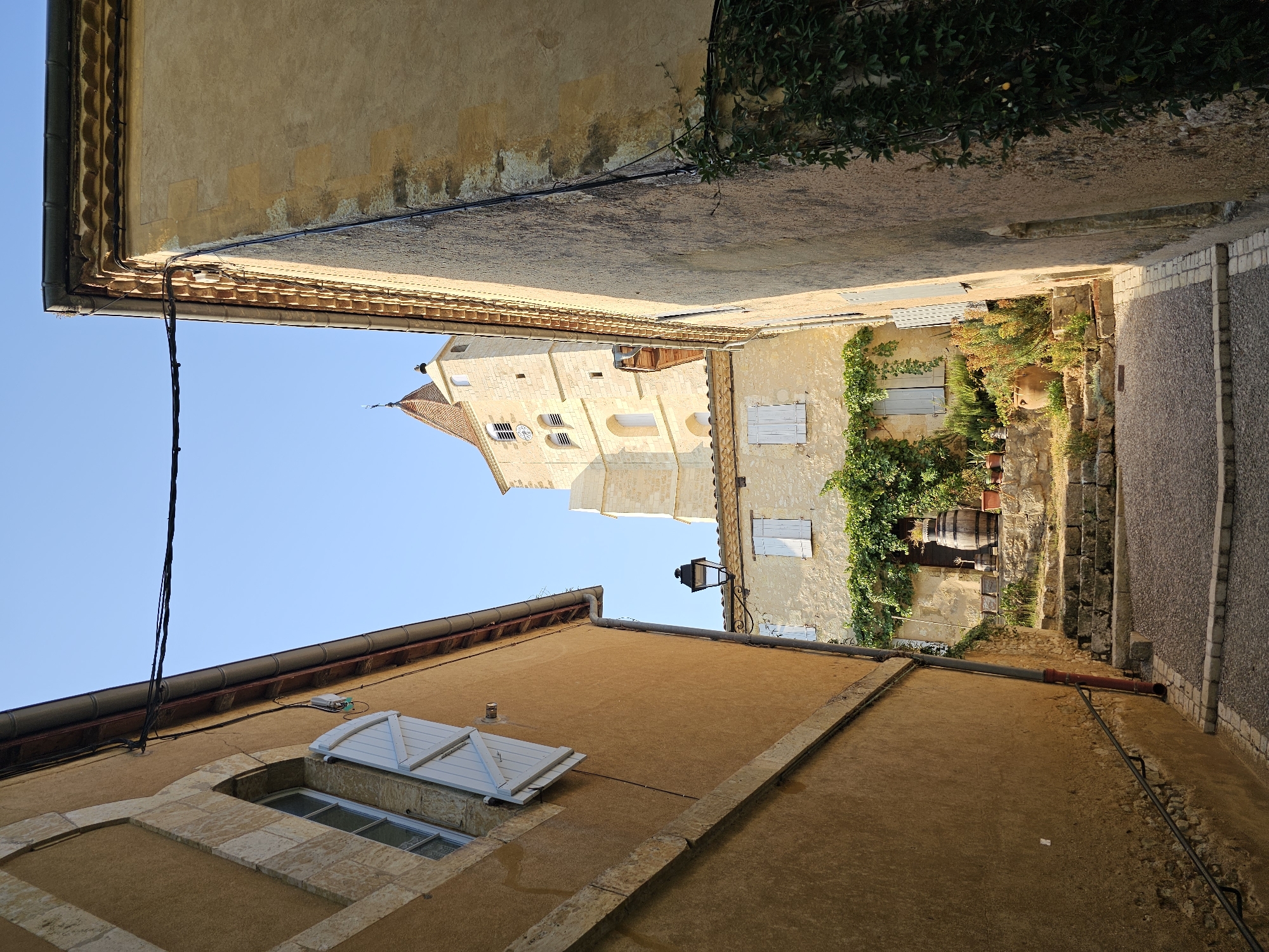The image shows a narrow alleyway in the village of Lavardens, France. The alley is paved with cobblestones and is lined with old stone buildings. In the background, a tall bell tower can be seen, which is part of the village church. The sky is clear and blue, and the sun is shining. There are no people visible in the image. The overall impression is one of peace and tranquility.