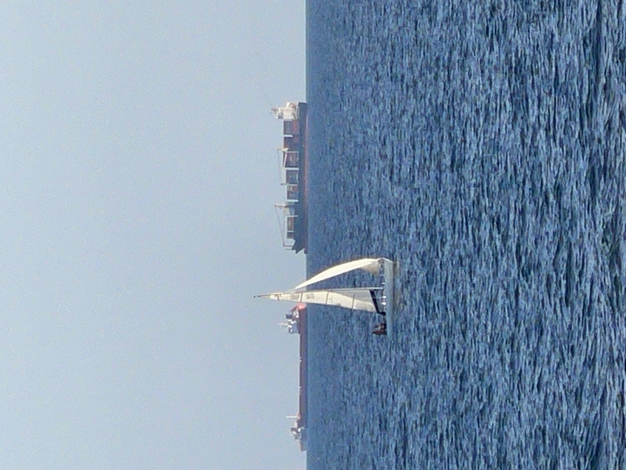 This media file shows a large cargo ship and a small sailboat on a body of water, likely the sea. The cargo ship, with its red hull and white superstructure, is positioned further away from the viewer. The sailboat, with white sails, is closer and appears to be sailing. The water is a deep blue and appears choppy, suggesting a breezy day. The sky is a clear, light blue, indicating fair weather. The overall scene depicts a maritime setting, possibly near a coast, given the presence of large cargo ships. The time of day appears to be daytime, likely late morning or early afternoon due to the lighting. There are no visible people or text in the image. The orientation of the image is vertical.