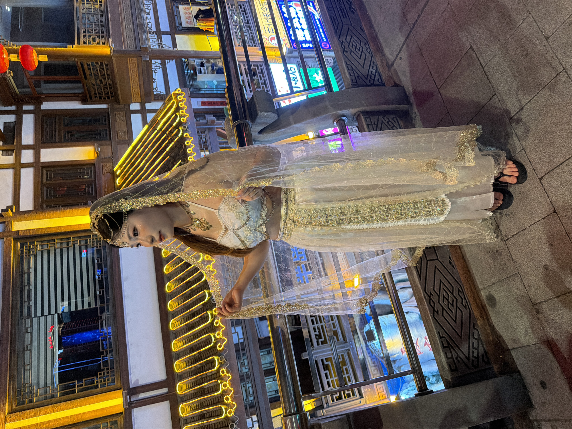A young woman, dressed in a white and gold outfit resembling traditional Indian attire with a veil, stands on a paved walkway at night. She is wearing a bejeweled headdress and necklace. Her attire consists of a crop top and a long skirt with intricate gold embroidery. She is holding the veil with her left hand. The background features a building with traditional Chinese architectural elements, illuminated by yellow lights. Red lanterns are visible hanging from the building. There are also neon signs with Chinese characters on other buildings further in the background. The walkway appears to be part of a street or pedestrian area in Yuzhong District, China, likely a tourist or shopping district given the illuminated signs and architecture. The lighting suggests it is evening or night.