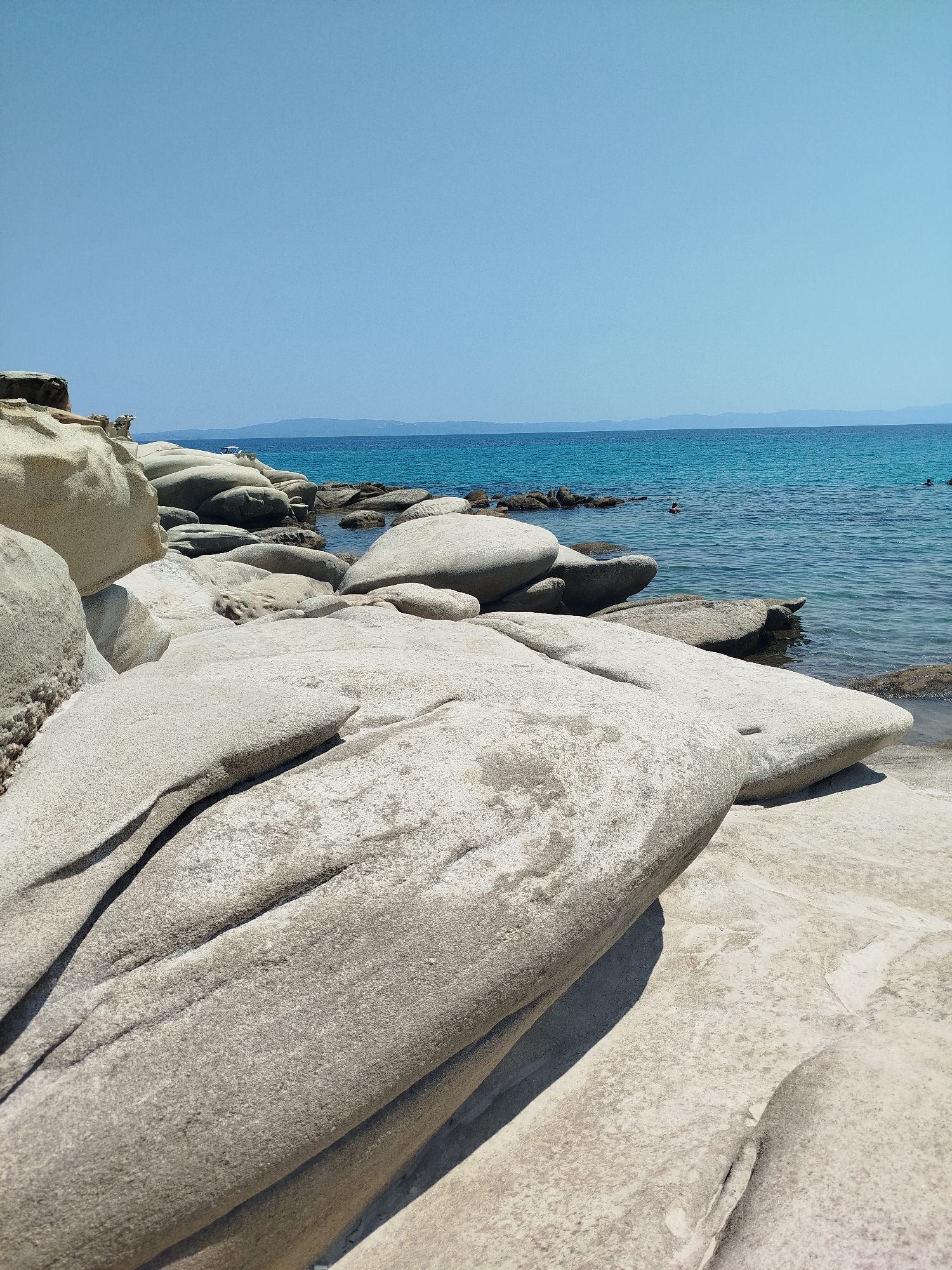 The image shows a rocky beach scene in Sithonia Municipal Unit, Greece. In the foreground, large, smooth, light-colored rocks dominate the landscape, with their textured surfaces illuminated by sunlight. These rocks are clustered together, creating natural formations and pathways leading towards the water.
The middle ground features more of these weathered rocks partially submerged in the clear, turquoise sea. A few darker, smaller rocks are visible along the waterline and in the water. The sea is calm with gentle ripples.
In the background, the blue expanse of the sea extends to the horizon, where a hazy silhouette of a distant coastline is visible. The sky is a clear, bright blue, with no clouds. Two people are faintly visible swimming in the sea in the distance. The overall impression is a serene and natural coastal environment.