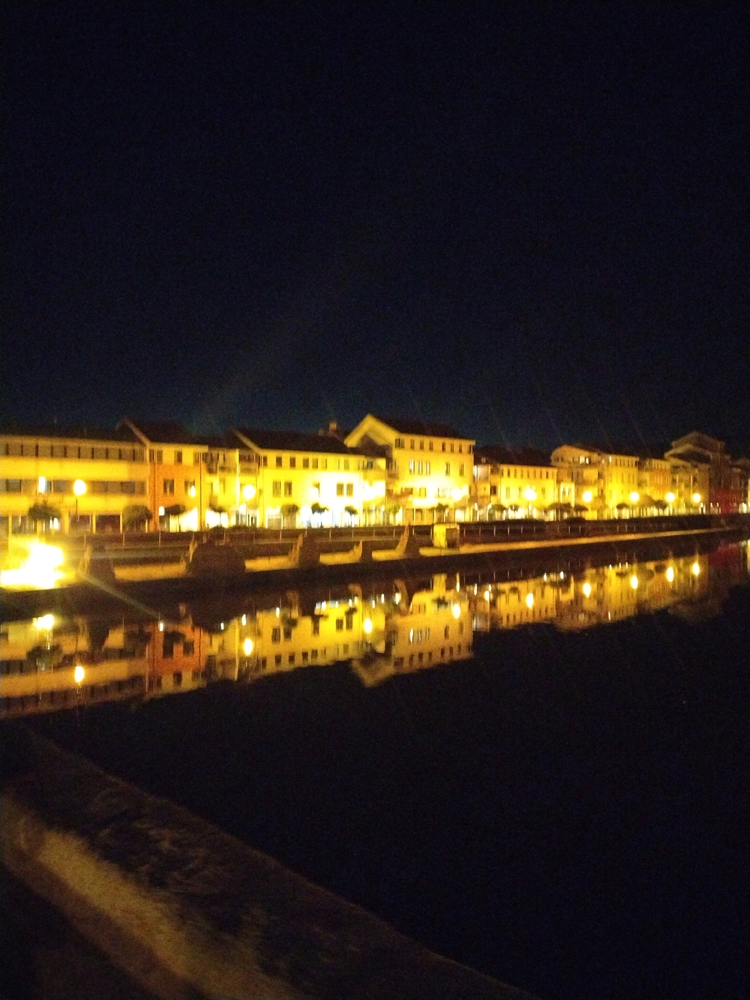 The image shows a night view of the Písek riverfront buildings illuminated by streetlights and interior lights. The buildings are a mix of architectural styles and are reflected in the dark water of the Otava River. A low stone embankment runs along the edge of the river in the foreground. The sky is dark and clear. There are no people visible in the image.