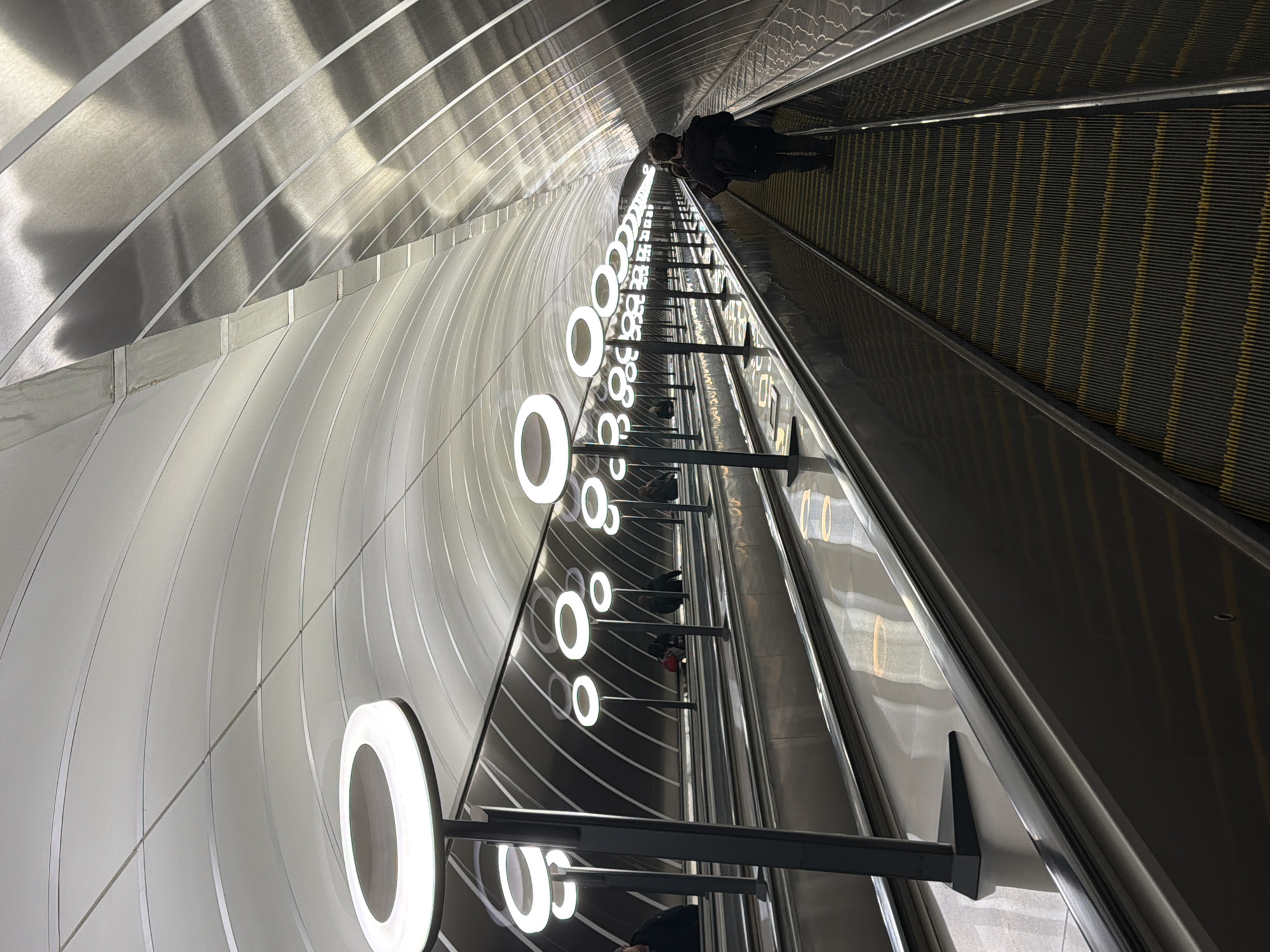 An upward-tilted shot captures a long, deep escalator tunnel, likely within a modern metro station in Moscow, Russia. The escalator, positioned on the right, features dark grey treads with prominent thin yellow horizontal lines, and black handrails. Several people in dark clothing are seen riding the escalator upwards, with one figure clearly visible towards the top of the frame.
The left wall of the tunnel is decorated with a series of striking, glowing white circular lights, mounted on dark vertical poles, which stretch far into the distance, creating a sense of immense depth. These lights illuminate the scene and are highly reflected on the curved, polished metallic walls and ceiling of the tunnel. The reflective surfaces create dynamic patterns of light and shadow, emphasizing the tunnel's curved architecture. The overall impression is one of modern design and functional infrastructure. The setting is indoors, so time of day and weather are not discernible. No legible text is visible within the image.