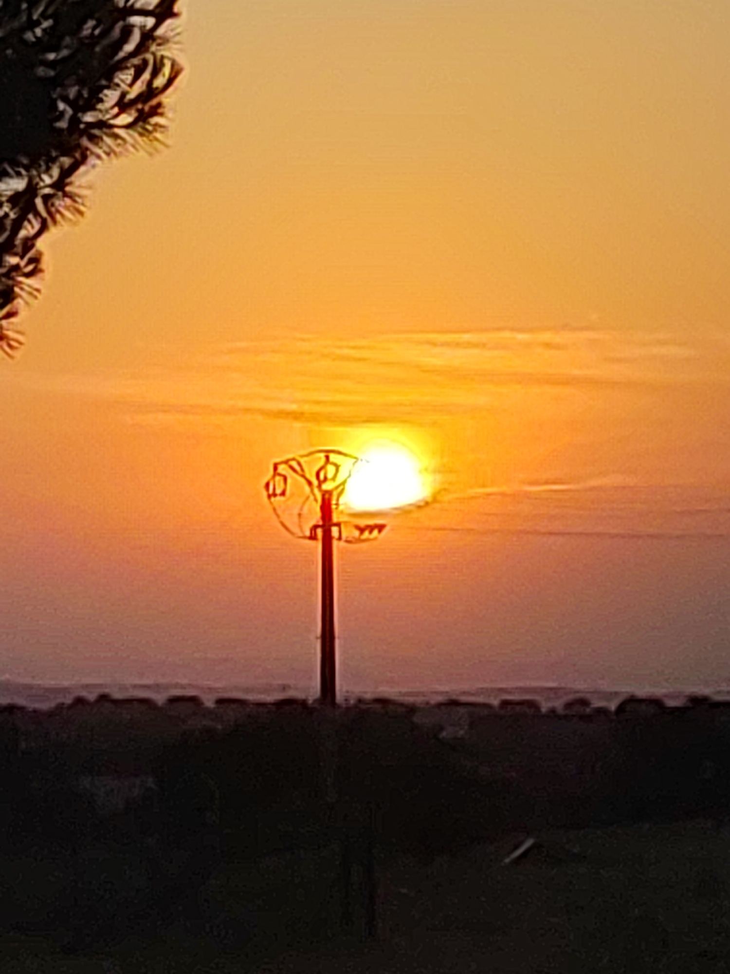 A street lamp, partially silhouetted against a setting sun, is visible in the foreground. The sun, a bright orange orb, is positioned slightly to the left of the lamp's center, casting a warm glow across the sky. The lamp has a decorative, possibly wrought-iron, top. The background consists of a dark, silhouetted landscape, likely trees or buildings, with a faint horizontal line of lighter color suggesting a distant horizon or body of water. The image appears to be taken in Sobradillo, Spain, judging by the context.
