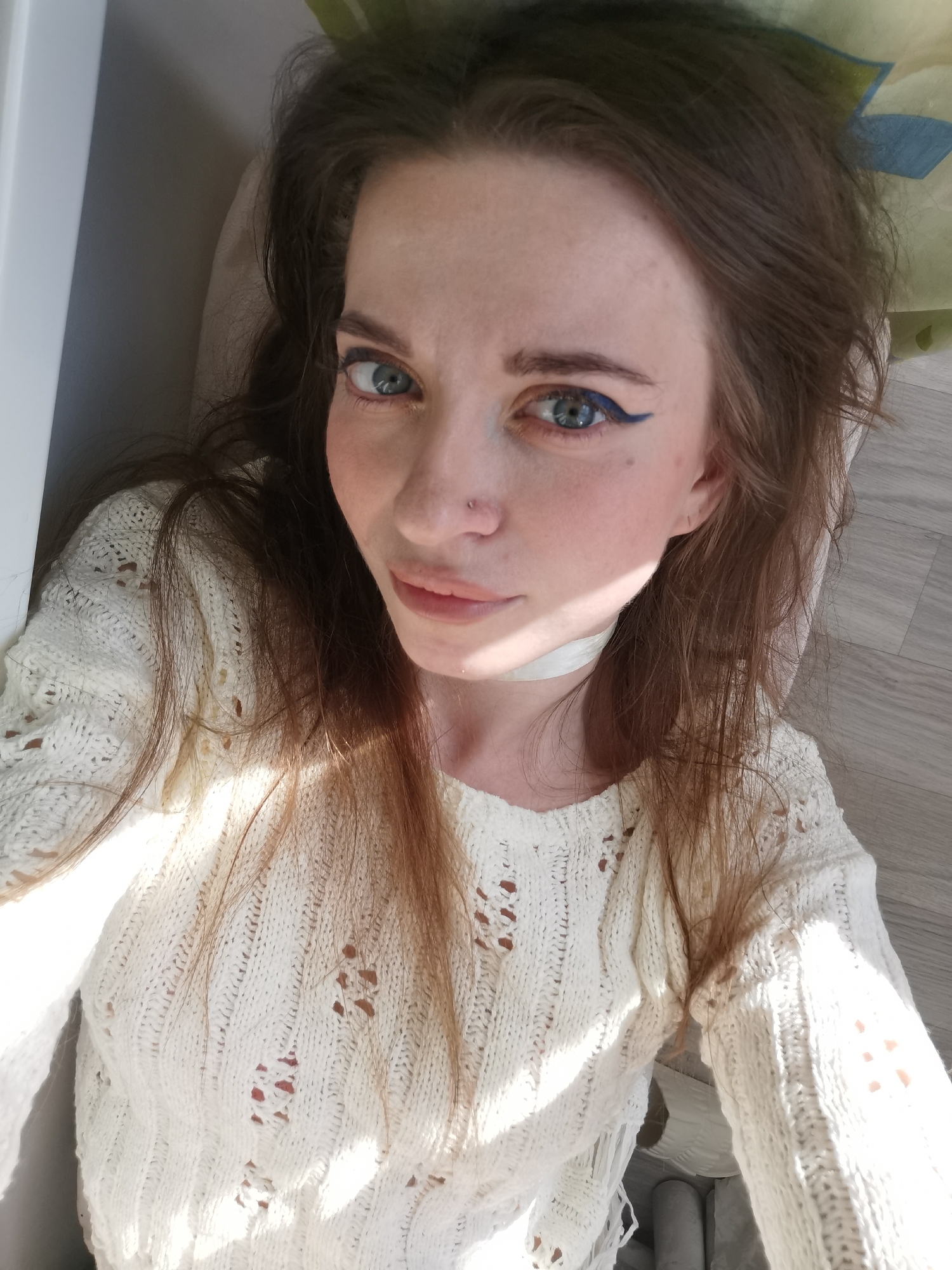 A woman is pictured in an indoor setting, likely a residence in Unknown, Unknown. She is wearing a cream-colored, knitted sweater with open-weave detailing and a white choker. Her hair is brown and loosely styled. She has blue eyes accented with blue eyeliner. She has a nose piercing. Her expression is neutral. The background consists of a white wall, a light-colored floor, and a partially visible green floral pattern.