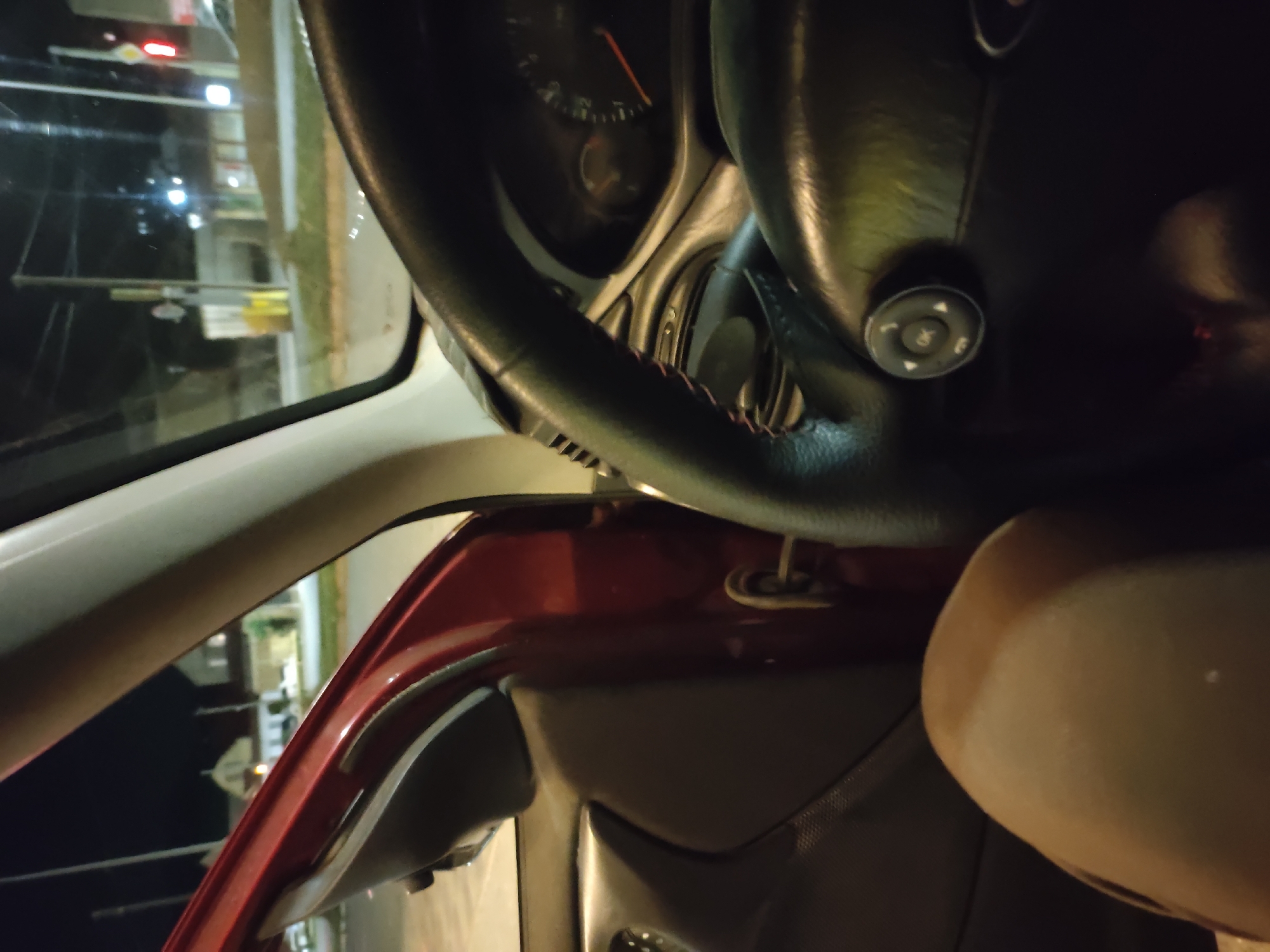 The image shows the interior of a car at night, with the driver's side door open. The interior is dark, with some ambient light coming from outside. The steering wheel and dashboard are visible, as is the driver's leg. Outside the car, a street with buildings and traffic lights can be seen. The scene appears to be in a residential area of Leninkent, Russia. There are no people or interactions visible.