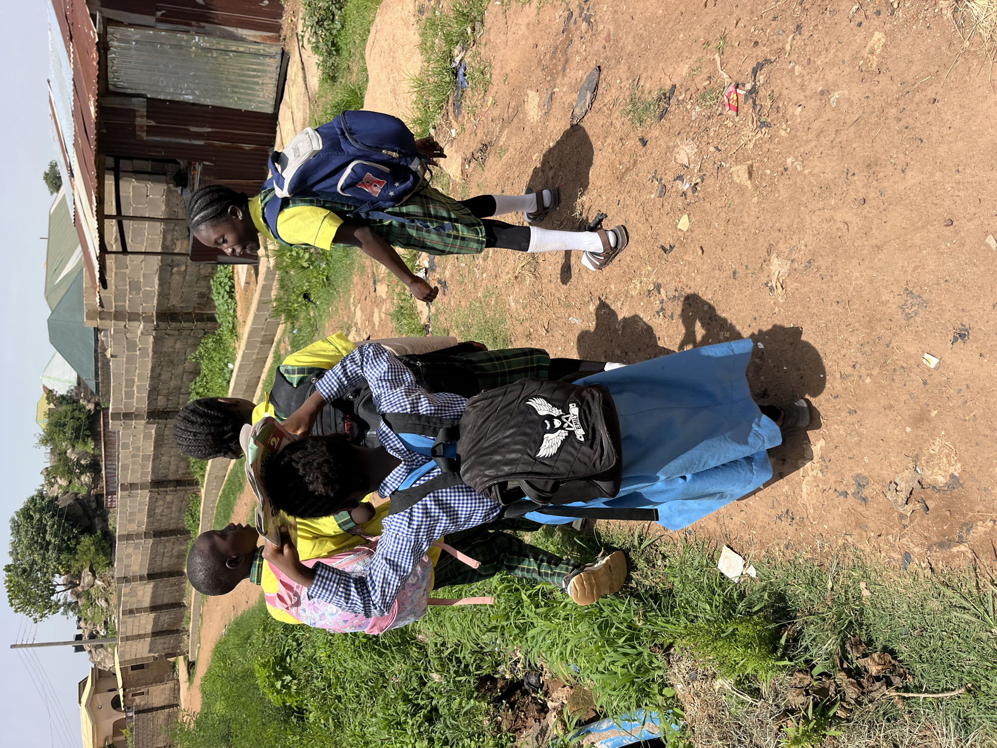 Four young girls in school uniforms are walking on a dirt path in an urban or semi-urban area. The girl in the front right wears a yellow shirt with plaid green and blue accents, a blue backpack, white socks, and brown sandals. The girl in the front left wears a blue and white checkered shirt, a pink backpack, and appears to be holding a book or papers. The girl behind her wears a yellow shirt and a black backpack. The girl in the back right is mostly obscured but appears to have on a yellow shirt and plaid clothing. The environment consists of dirt, sparse vegetation, a brick wall, a corrugated metal structure, and some scattered debris. The sky is visible and appears clear.