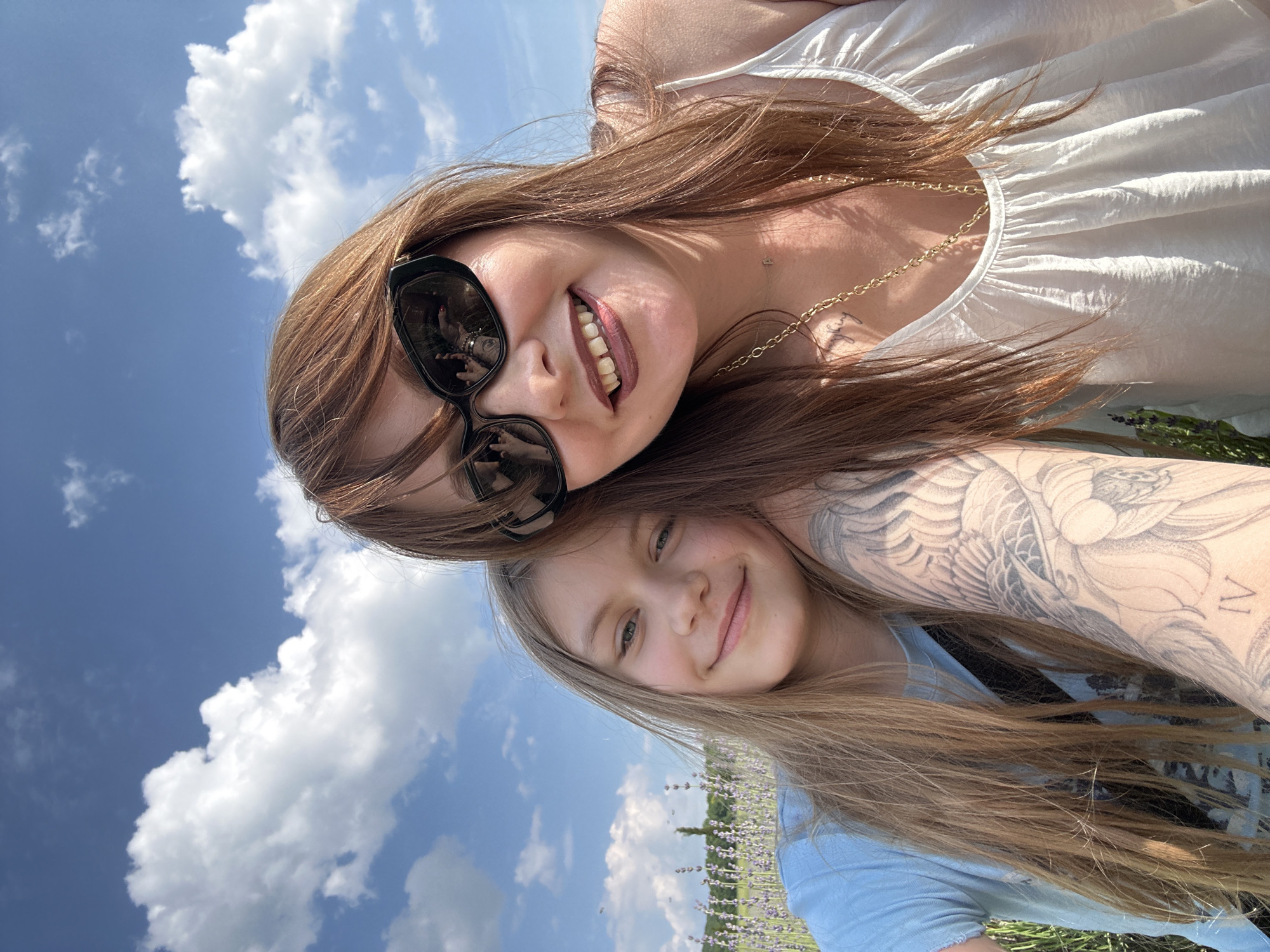 Two individuals, a woman and a younger person, are taking a selfie outdoors in a field of lavender. The woman, in the foreground on the right, wears sunglasses and a white top with a gold necklace. She has auburn hair and a tattoo on her left arm. The younger person, on the left, has long blonde hair and wears a light blue t-shirt. They are both smiling and appear to be posing for the photo. The background shows a clear blue sky with white clouds and rows of purple lavender plants. This scene is consistent with the common tourist activities and landscapes found in Pavlovskiy, Russia, particularly during the lavender blooming season.