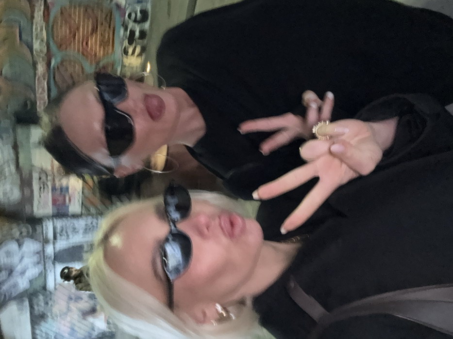The image depicts two female individuals posing for a photograph, likely a selfie, within the Leake Street Arches graffiti tunnel in Lambeth, London, United Kingdom.
The individual on the left has short, blonde hair and wears dark, oval-shaped sunglasses, multiple small hoop earrings, and a dark top. She has her lips pursed and makes a "peace" or V-sign with her left hand, displaying a prominent decorative ring on her right ring finger.
The individual on the right has dark hair pulled back and wears large, dark sunglasses and large gold hoop earrings. She is extending her tongue in a playful expression. Her right hand is partially visible, also making a V-sign.
The background features walls and archways completely covered in diverse, colorful graffiti and street art, characteristic of the Leake Street Arches environment. A blurred figure, appearing to be a person in a dark hat or helmet, is visible in the distant left background. The scene is lit by ambient light within the covered urban space.