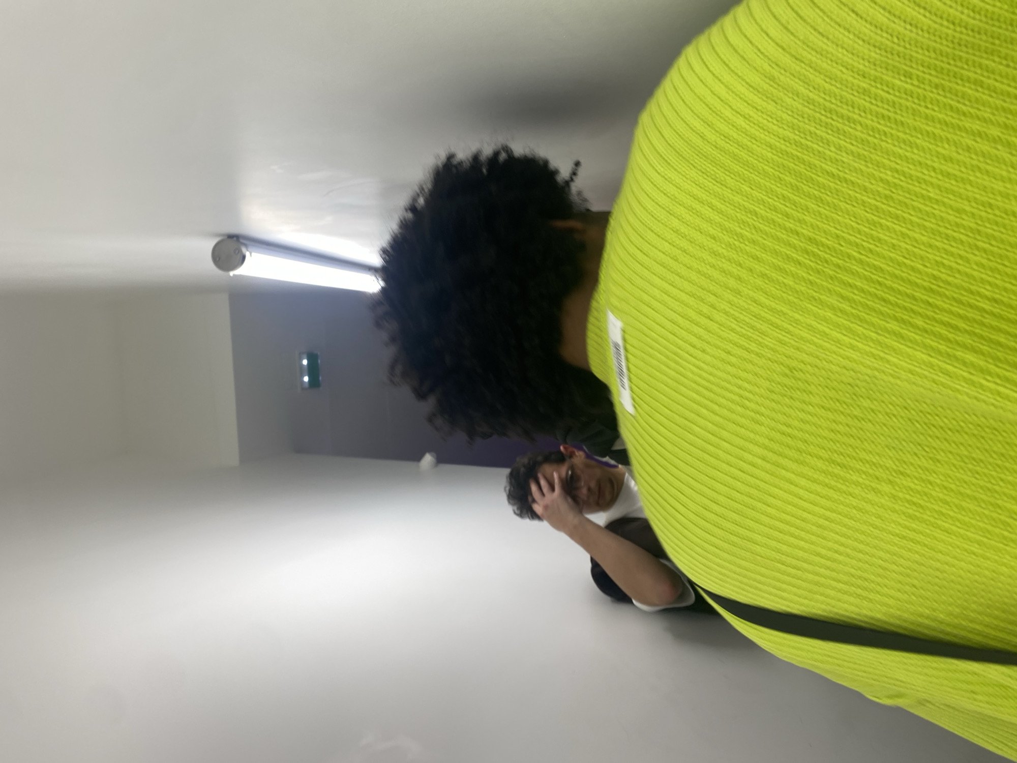 The image depicts an interior space with two individuals present. A person with dark curly hair is positioned in the foreground, partially obscured by a bright neon green knitted garment. Behind them, a second person is visible, appearing to hold their head with one hand. The background features a white wall, with a fluorescent light fixture mounted on the ceiling and a darker colored wall further back, possibly purple. This area includes a small window or opening. The scene gives the impression of a studio or gallery space due to its minimalist design and lighting. The location in Paris, France, cannot be confirmed based on visual information alone.