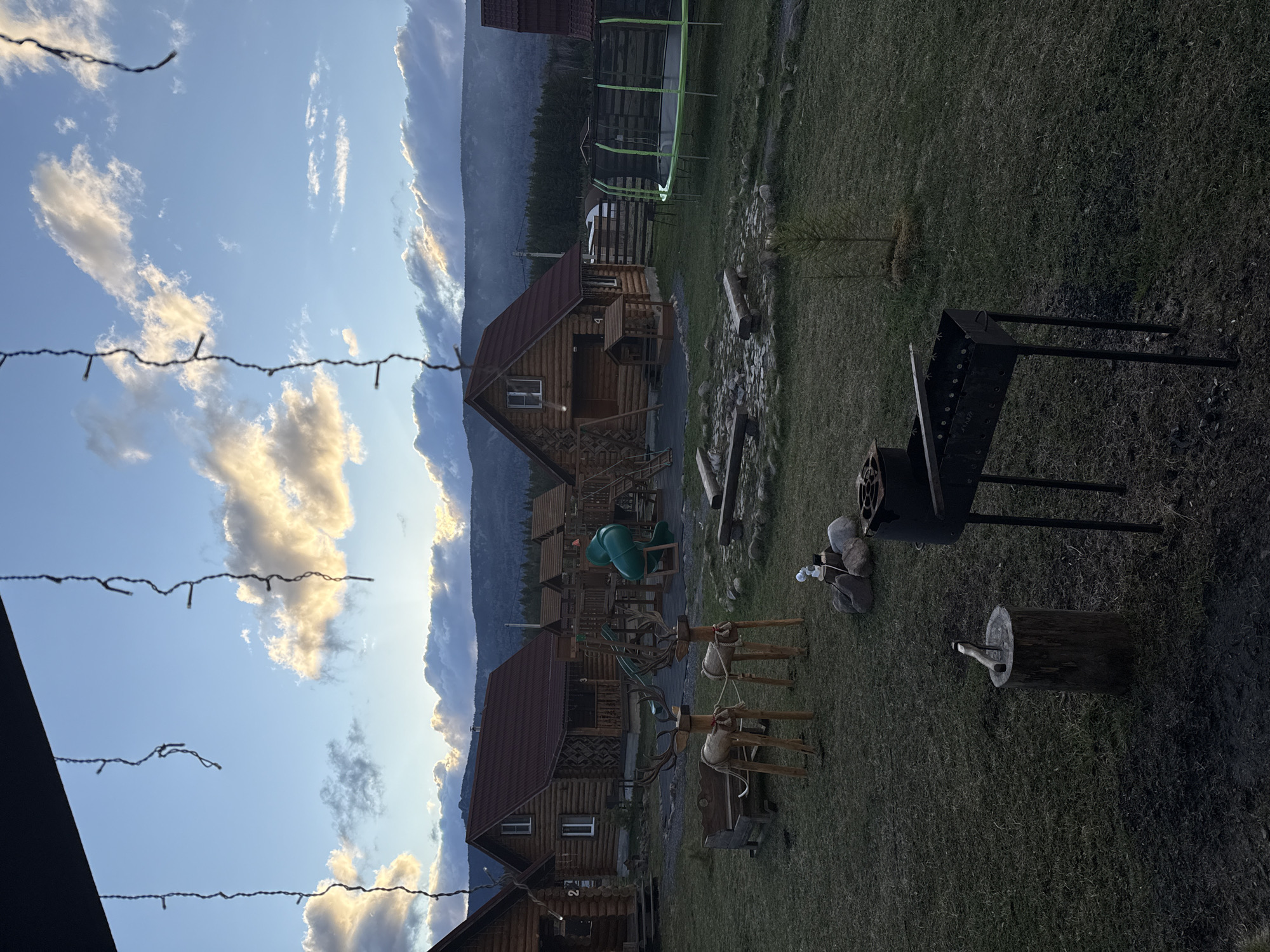 The image depicts an outdoor scene at dusk, likely in a rural or mountainous area, possibly in the city of Tyulyuk, Russia. Wooden cabins with dark red roofs are visible, suggesting a tourist or recreational resort. In the foreground, there are wooden deer statues, a grill, a log with an axe, and a small makeshift fire pit. A trampoline is visible behind one of the cabins. The sky is a mix of blue and clouds tinged with the setting sun's light, with mountains in the background. Strings of lights are strung across the top of the frame. The scene appears to be quiet and peaceful.