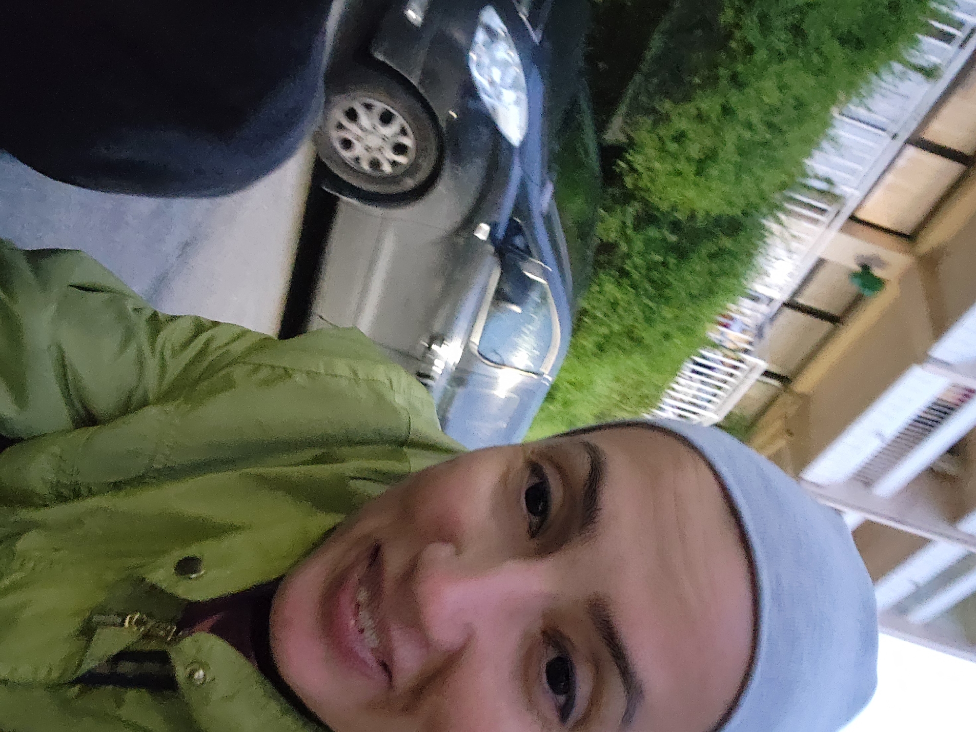 A selfie of a woman with short brown hair and a gray head covering, wearing a lime green jacket, is angled upwards. She is smiling at the camera. Behind her is a dark gray car with a visible wheel. Further back is green foliage that might be shrubbery or trees. In the upper right, there is part of a building with what appears to be a white metal railing or structure, hinting at a balcony. The environment suggests an outdoor setting, possibly in a residential or urban area. The location in Sochi, Russia is deduced from background information, but this is not directly discernible from the image.